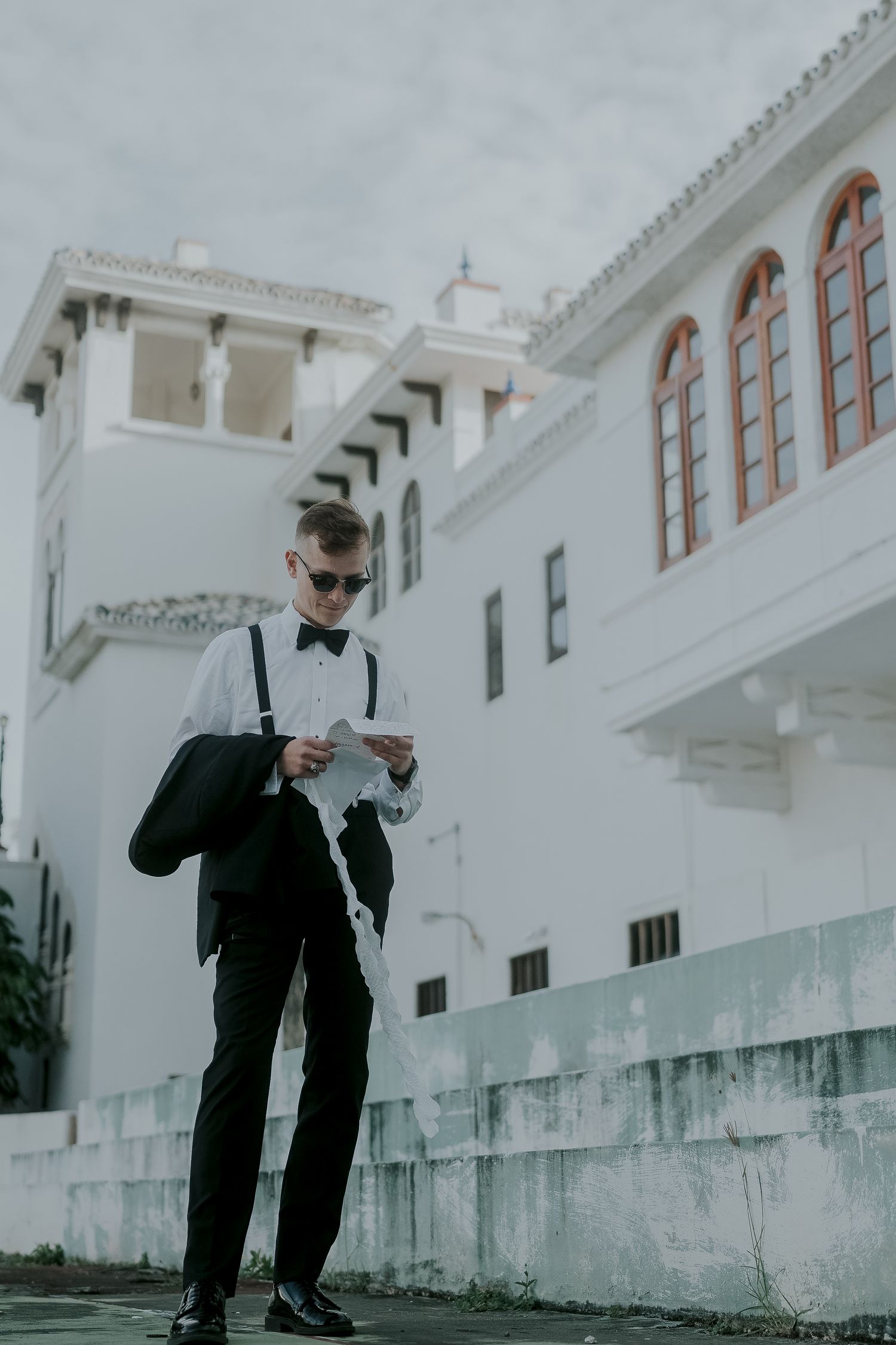 Groom in black tuxedo with suspenders and sunglasses reading outside a white colonial building.
