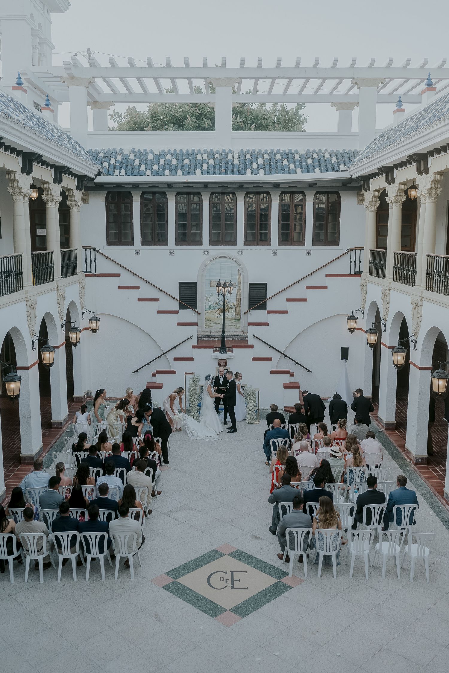Aerial view of elegant outdoor wedding ceremony in white Spanish courtyard with arched balconies and monogram floor.