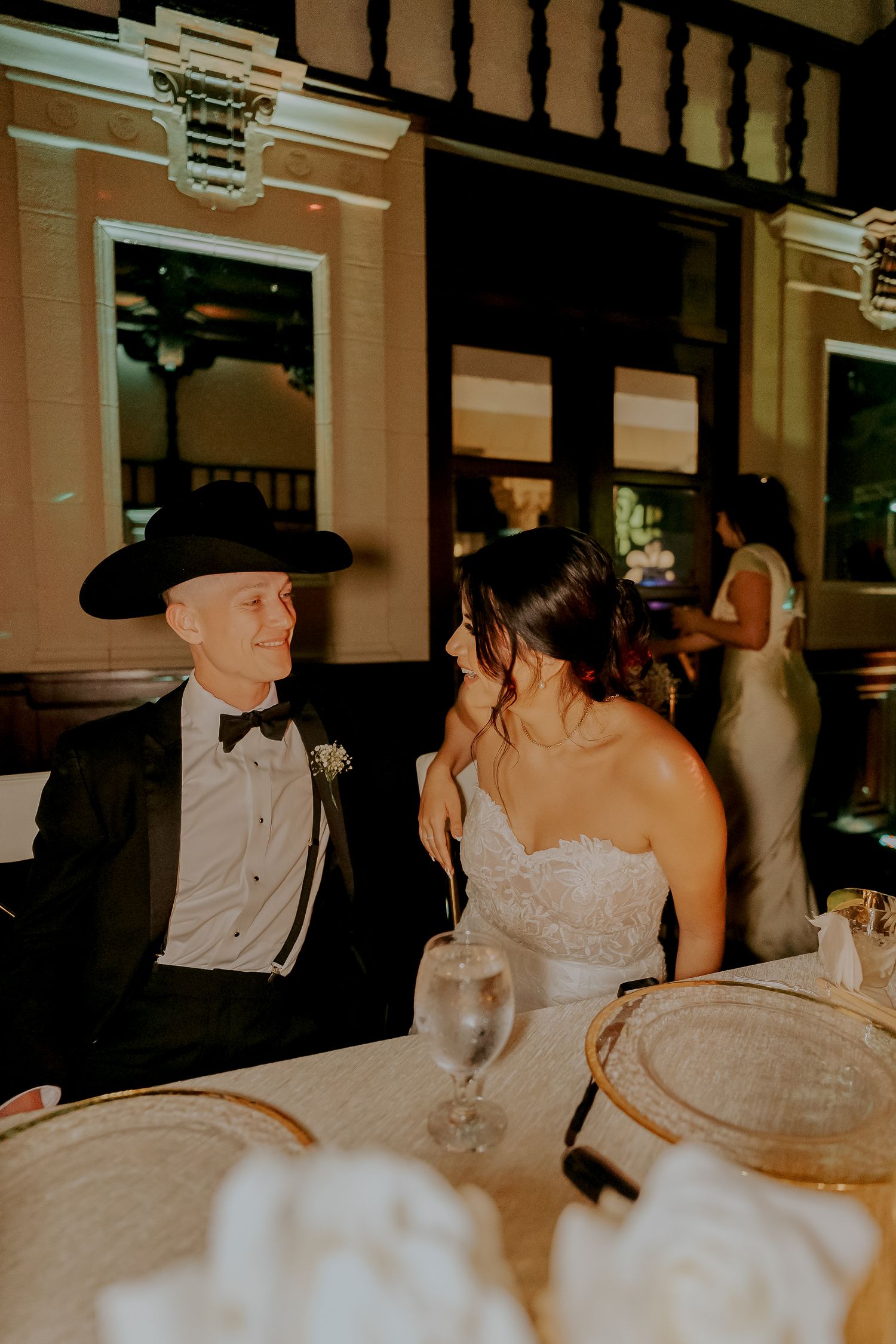 Groom in black cowboy hat and tuxedo smiles at bride during romantic wedding reception dinner.