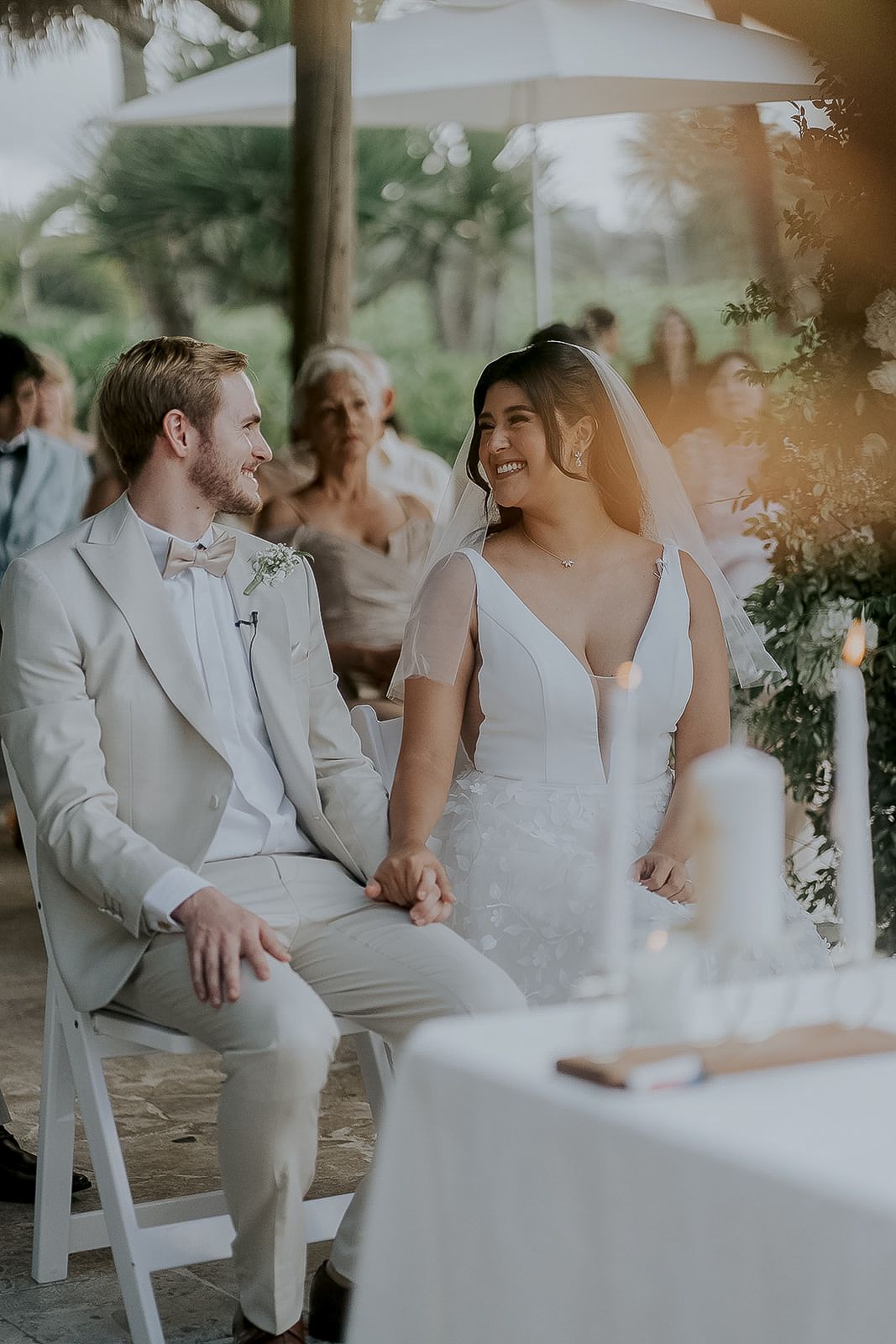 Bride and groom hold hands and smile during outdoor wedding ceremony, groom in beige suit, bride in white gown.