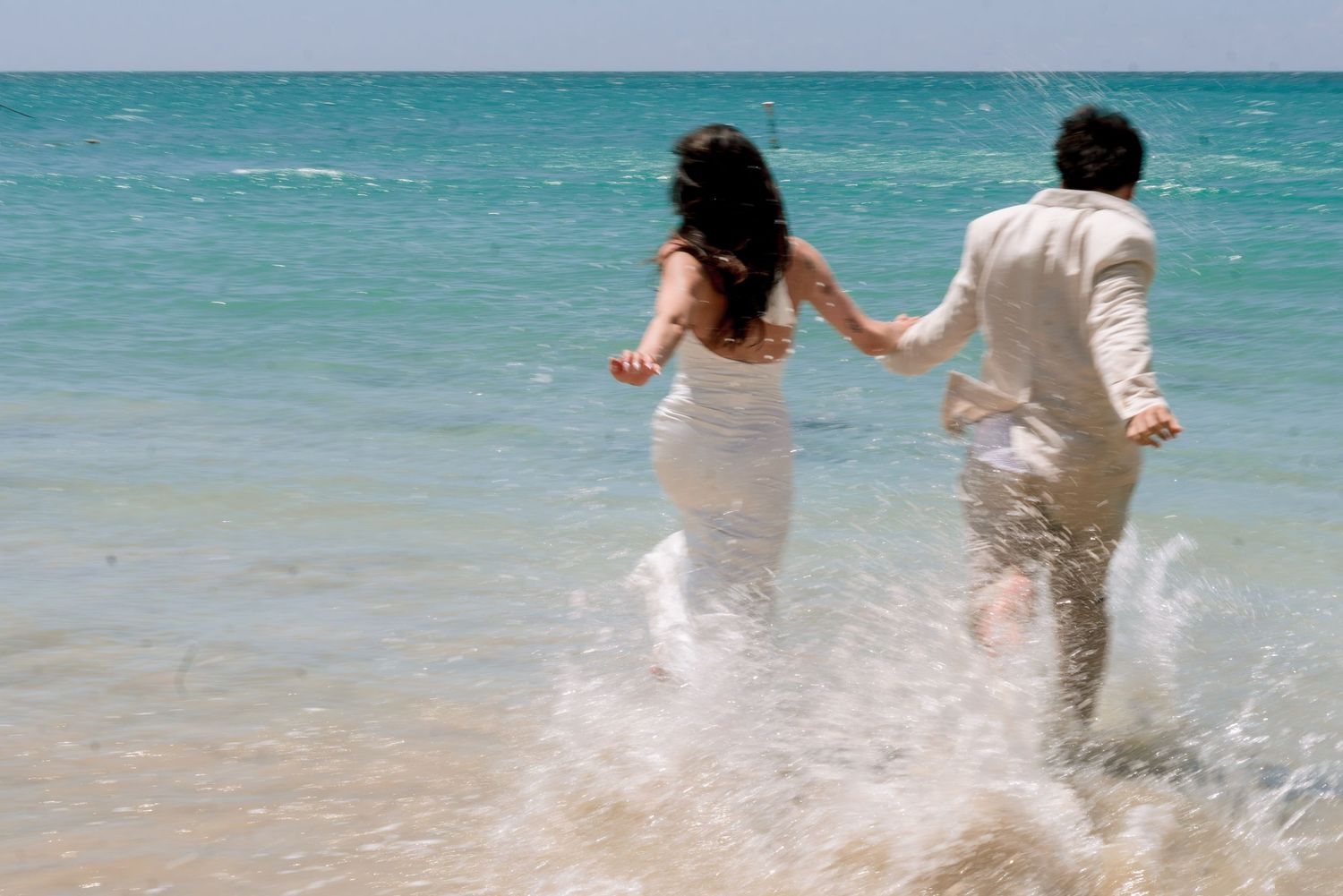 Newlywed couple in wedding attire running hand-in-hand through ocean waves on a sunny tropical beach.