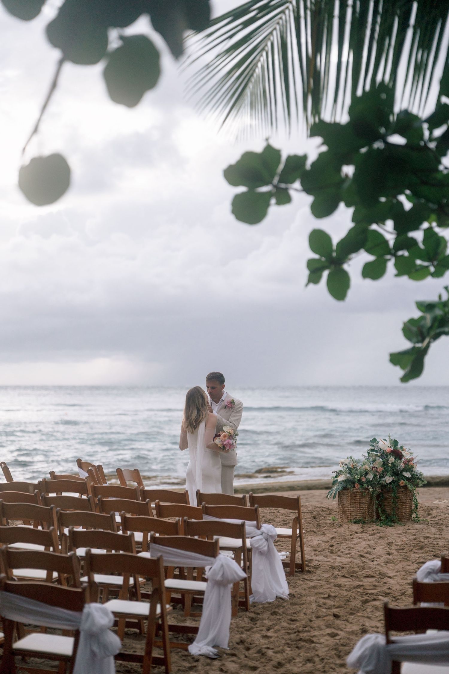 Couple sharing a romantic moment at a beach wedding ceremony with wooden chairs and ocean backdrop.