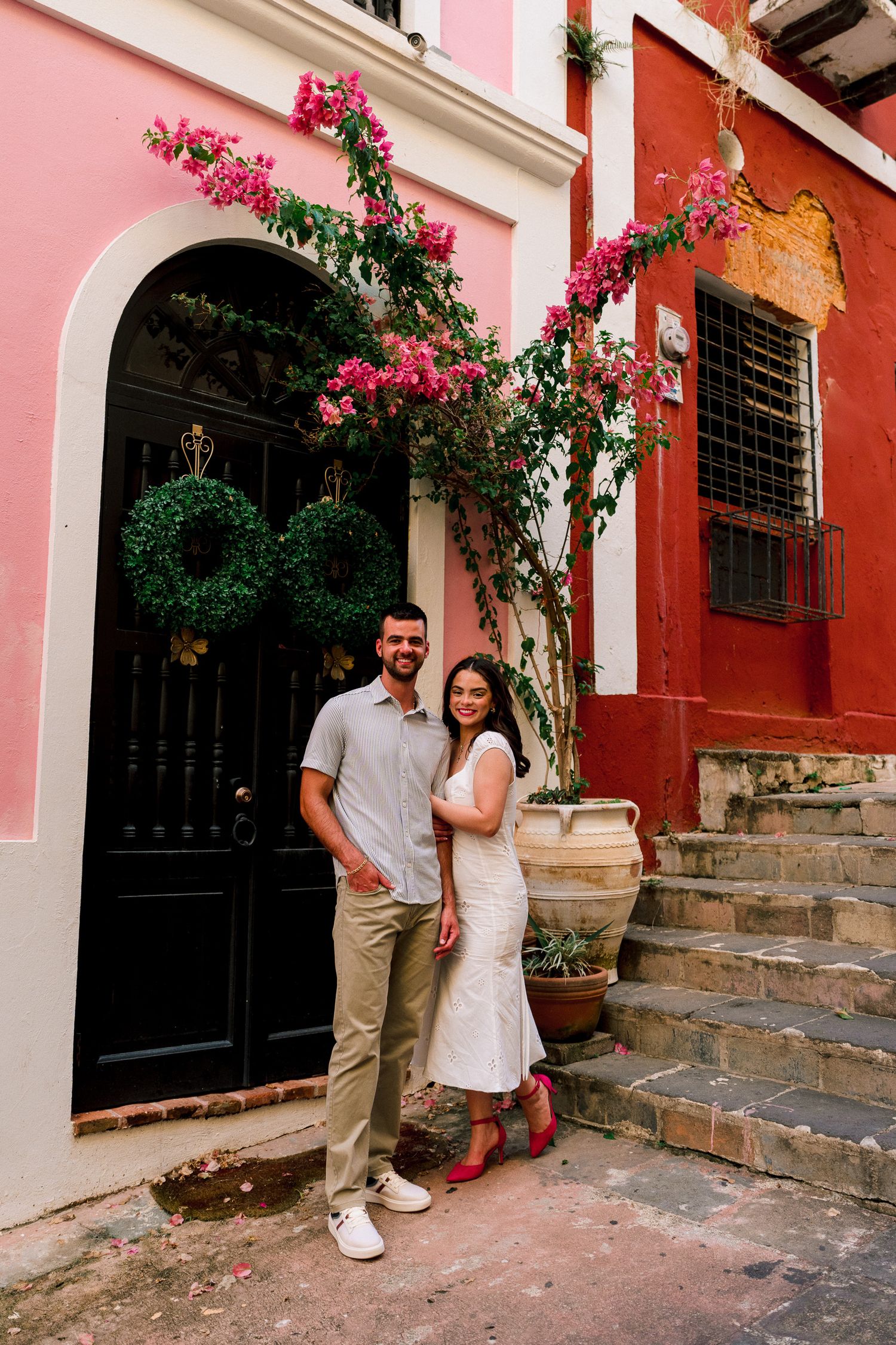 Couple smiling in front of colorful pink and red building with bougainvillea flowers and dark arched door.
