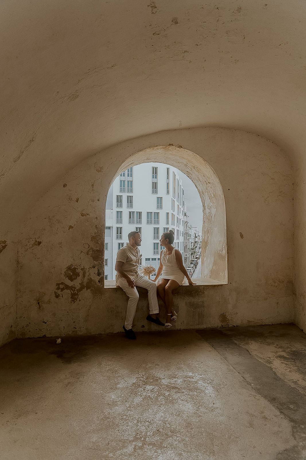 Couple sitting together in an arched stone window alcove with a city building visible outside.
