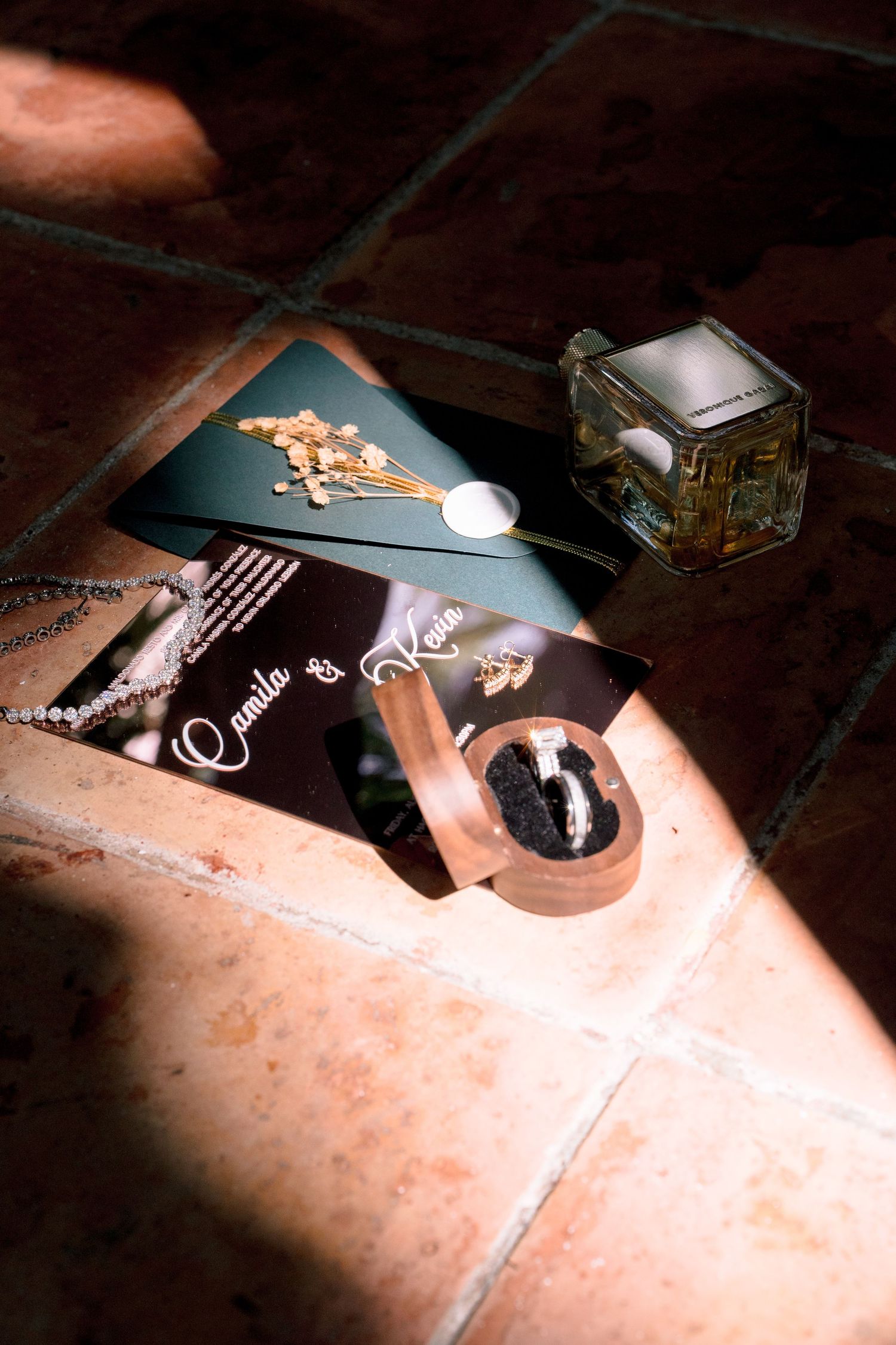 Wedding stationery with rings, perfume bottle, and dried flowers on terracotta tiles in warm sunlight.