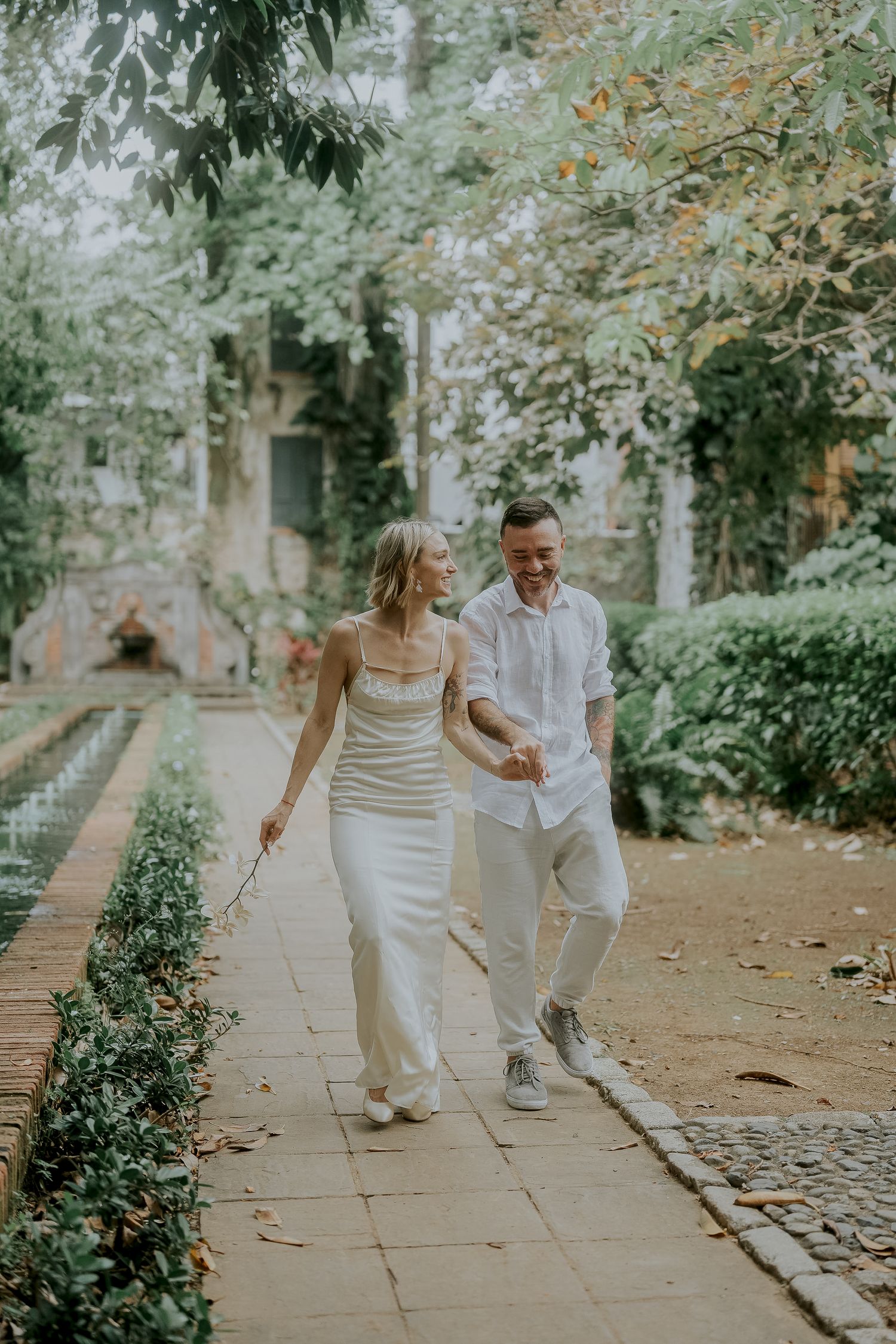 Happy couple holding hands walking along a garden path with green hedges and historic building backdrop.