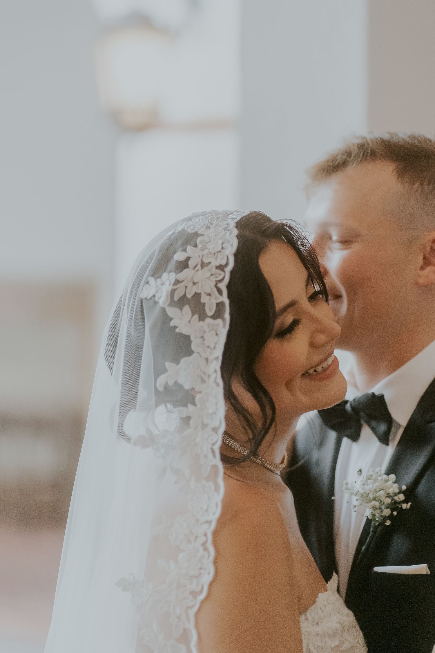 Groom kissing bride's cheek as she smiles, wearing lace veil and pearl necklace at wedding ceremony.