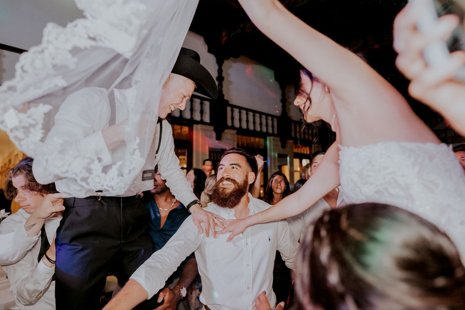 Groom dances joyfully under lifted wedding dress veil surrounded by cheering guests at reception.