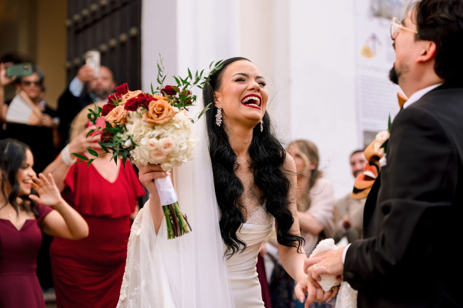 Joyful bride in white gown holding red and white bouquet laughing with groom at outdoor wedding ceremony.