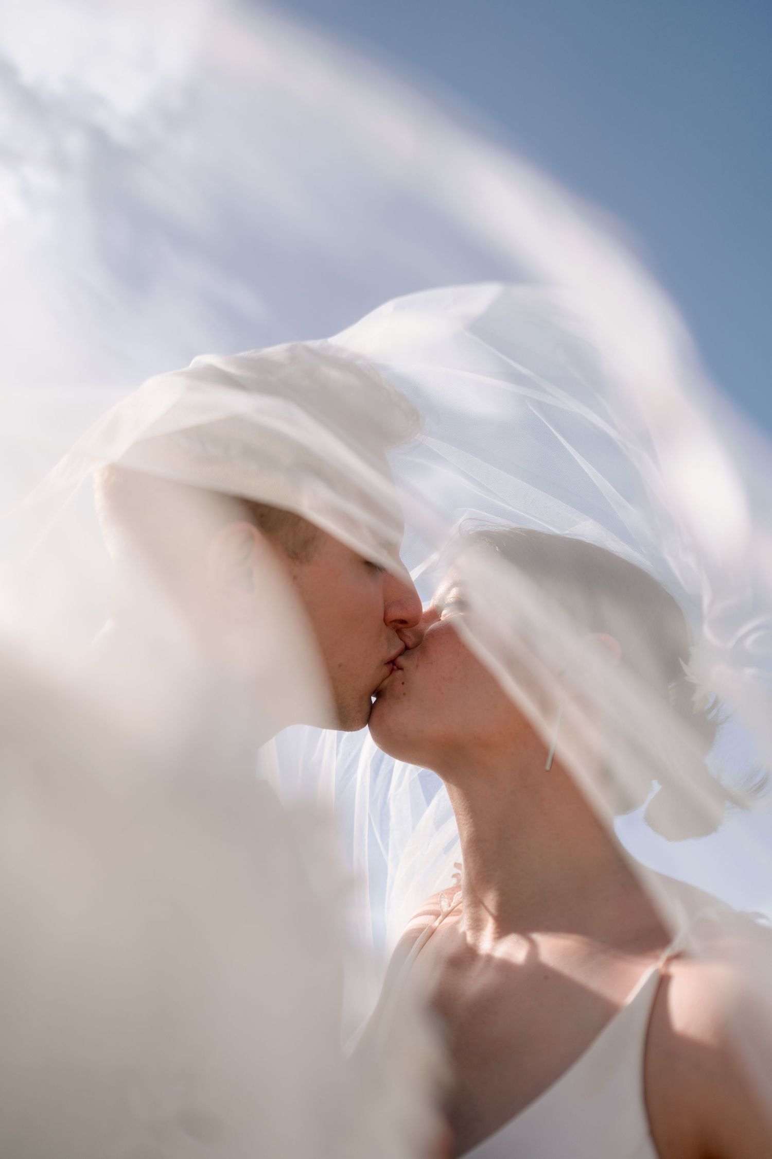 A bride and groom share a romantic kiss beneath a flowing white veil against a soft blue sky.
