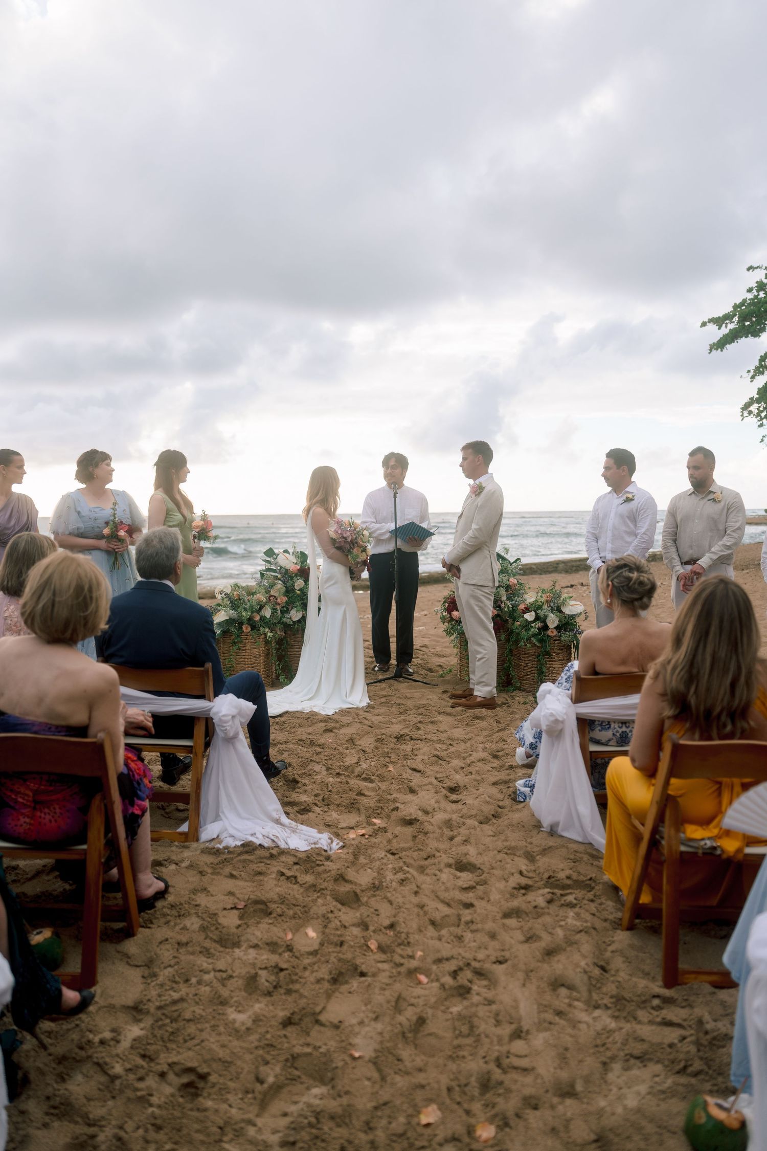 Romantic beach wedding ceremony with bride and groom at ocean altar, guests seated on sandy aisle under cloudy sky.