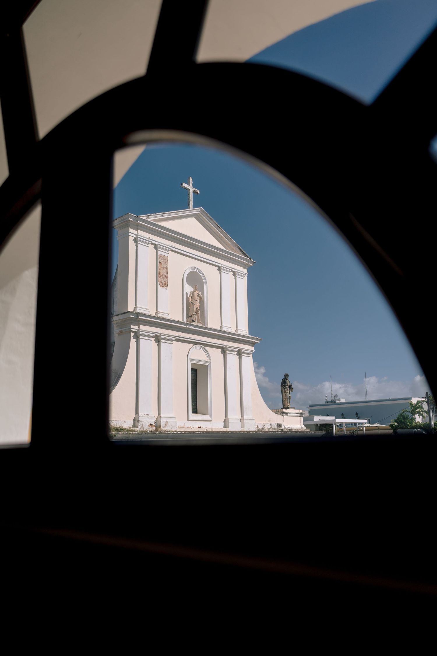 White colonial church facade with cross viewed through ornate iron gate arch against blue sky.