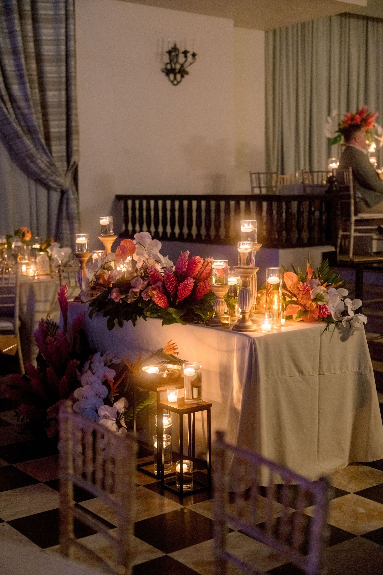 Romantic wedding sweetheart table adorned with tropical floral arrangements and glowing candles in lanterns.