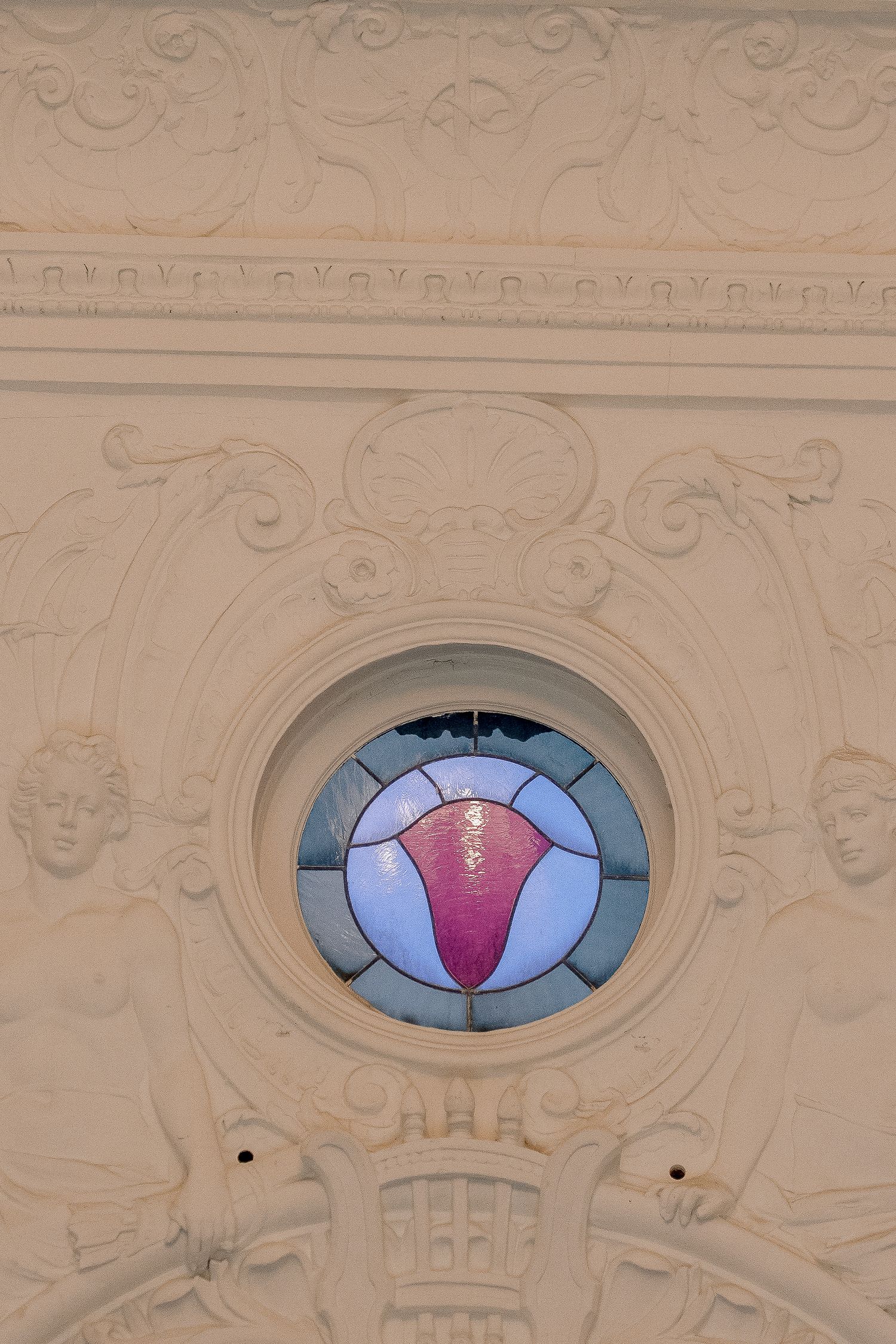 Ornate circular stained glass window with pink and blue design surrounded by white plaster relief sculptures.