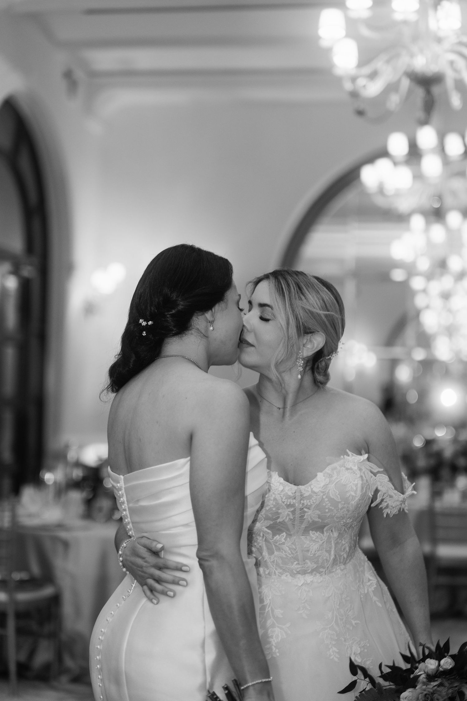 Two brides in white gowns share a tender kiss at their elegant wedding reception in black and white photo.
