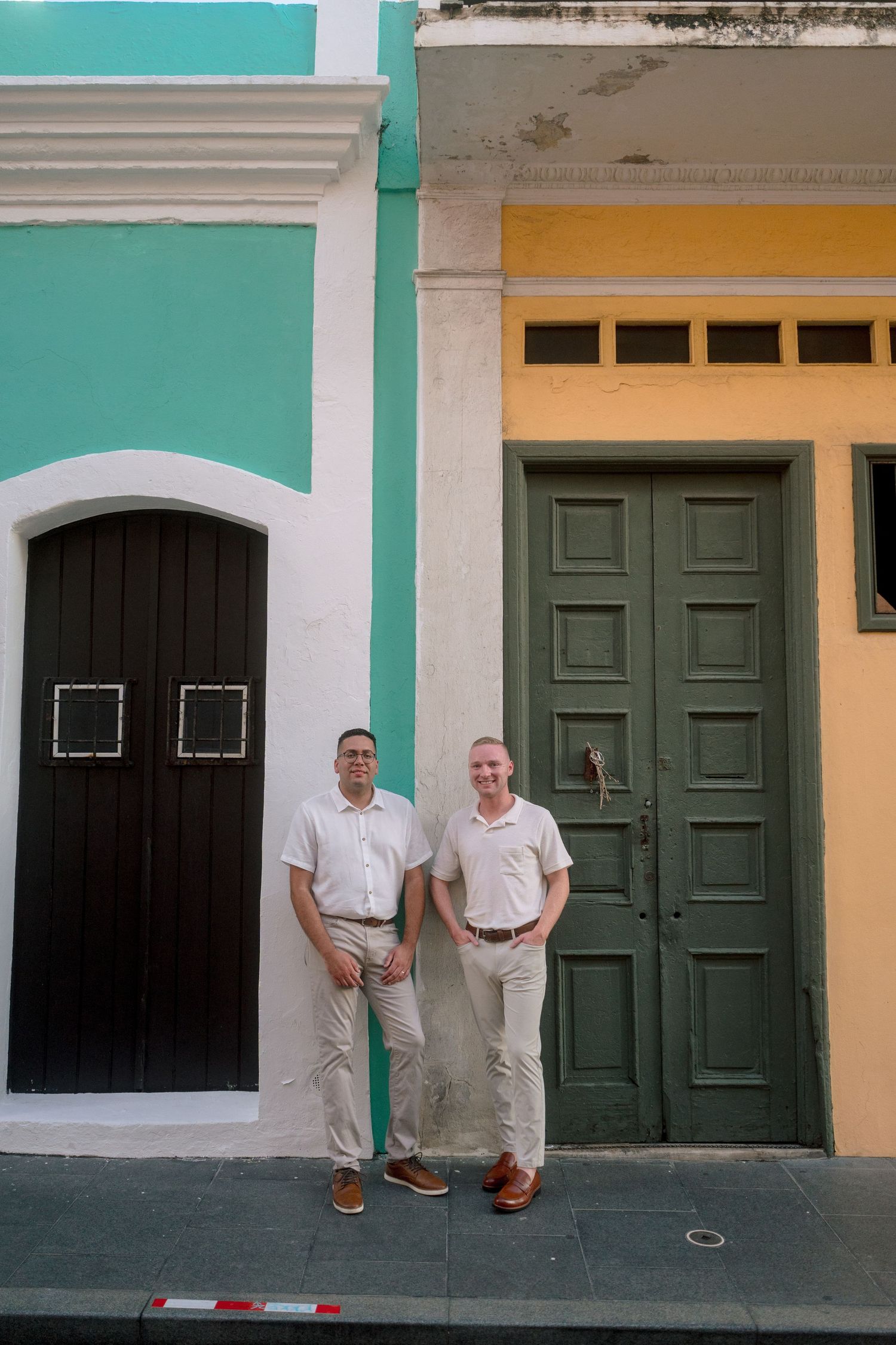 Two men in matching white shirts and khaki pants stand between colorful teal and yellow colonial buildings.