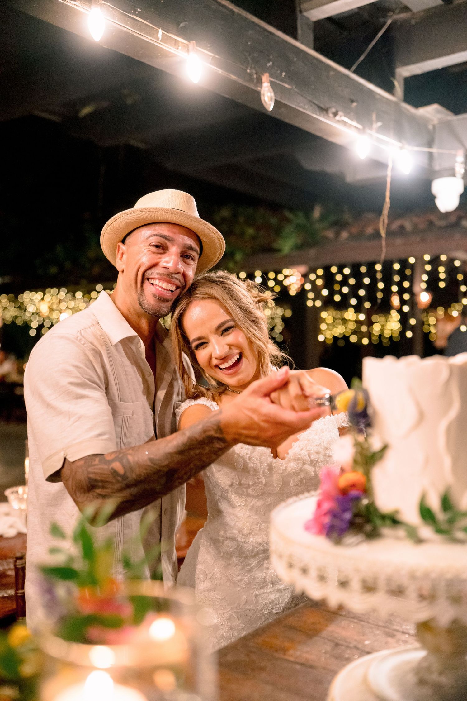 A smiling couple cuts their white wedding cake together at an outdoor evening reception with warm string lights in the background.