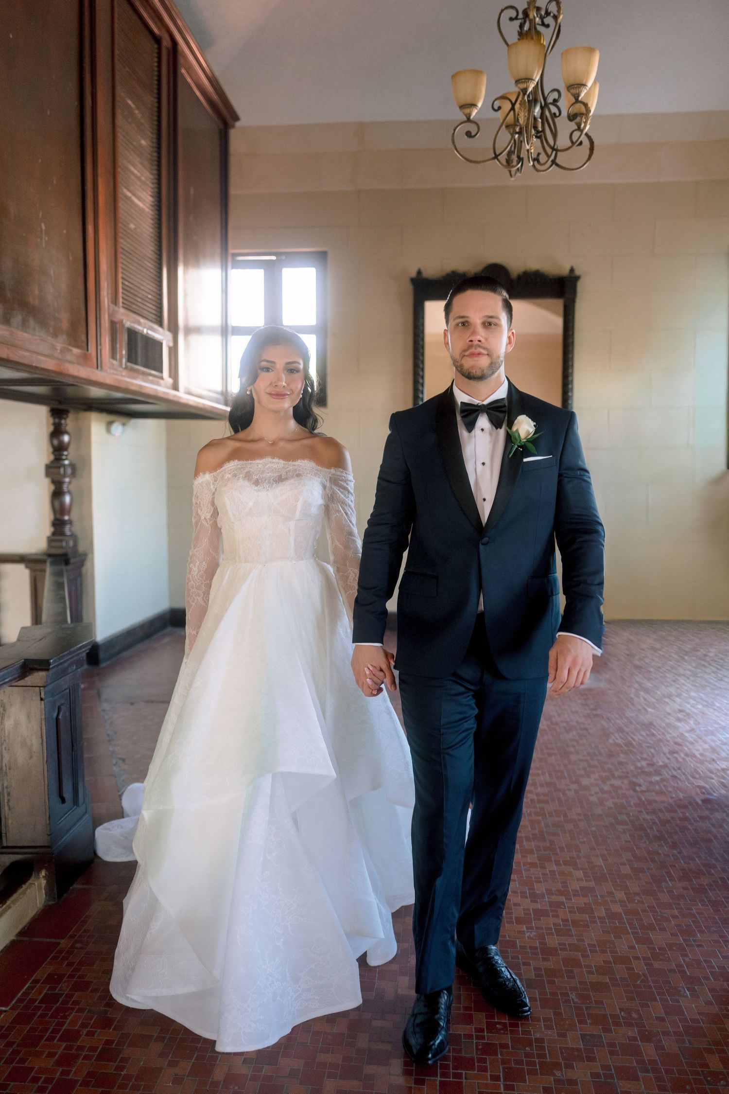 Bride in off-shoulder white gown and groom in black tuxedo hold hands walking through elegant venue.