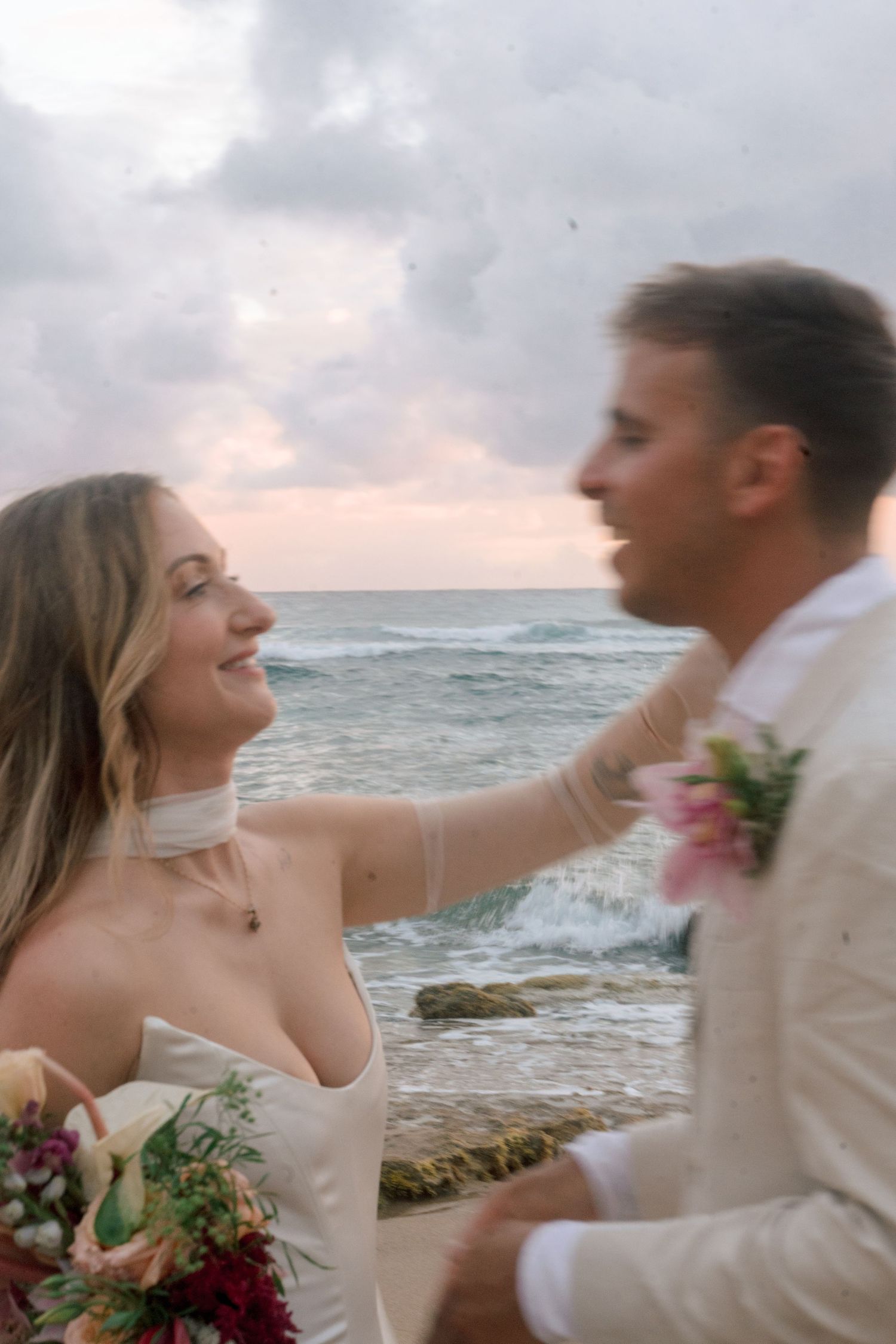 Bride in white gown tenderly touches grooms face while smiling with ocean waves crashing behind them.