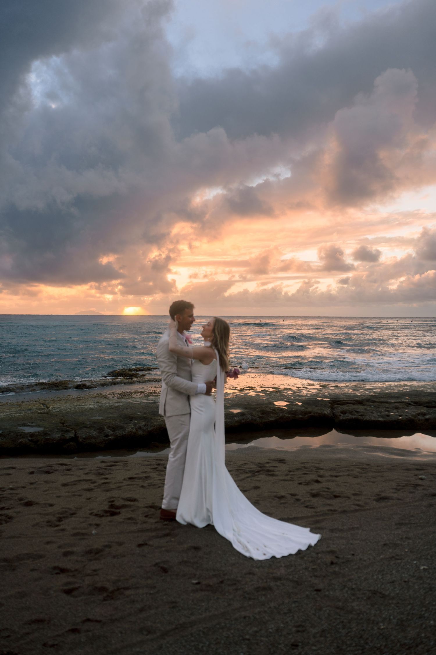 Bride and groom embrace romantically on beach rocks during stunning pink and purple sunset after wedding.