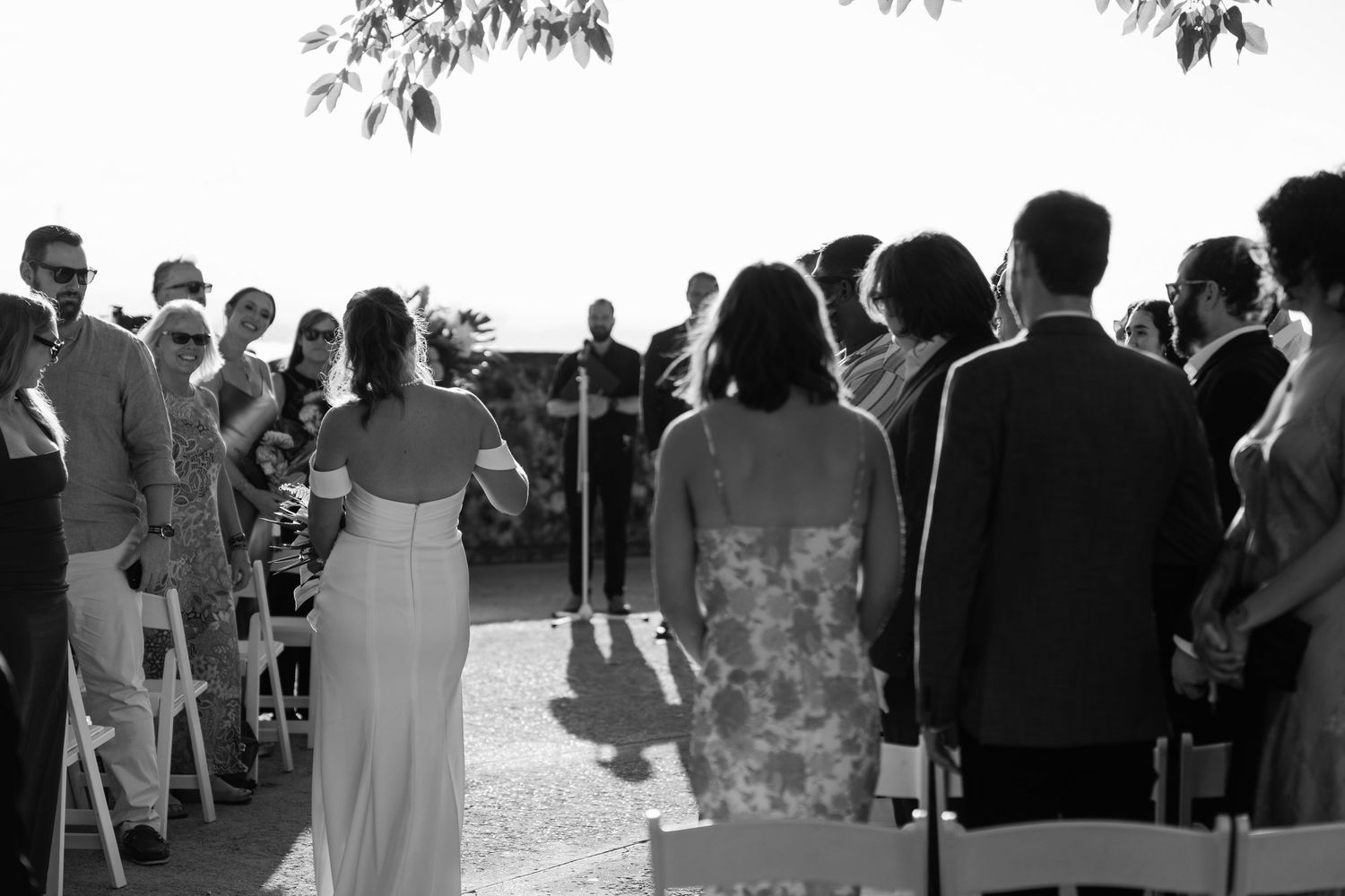 Black and white photo of outdoor wedding ceremony with guests seated and standing in sunlight.