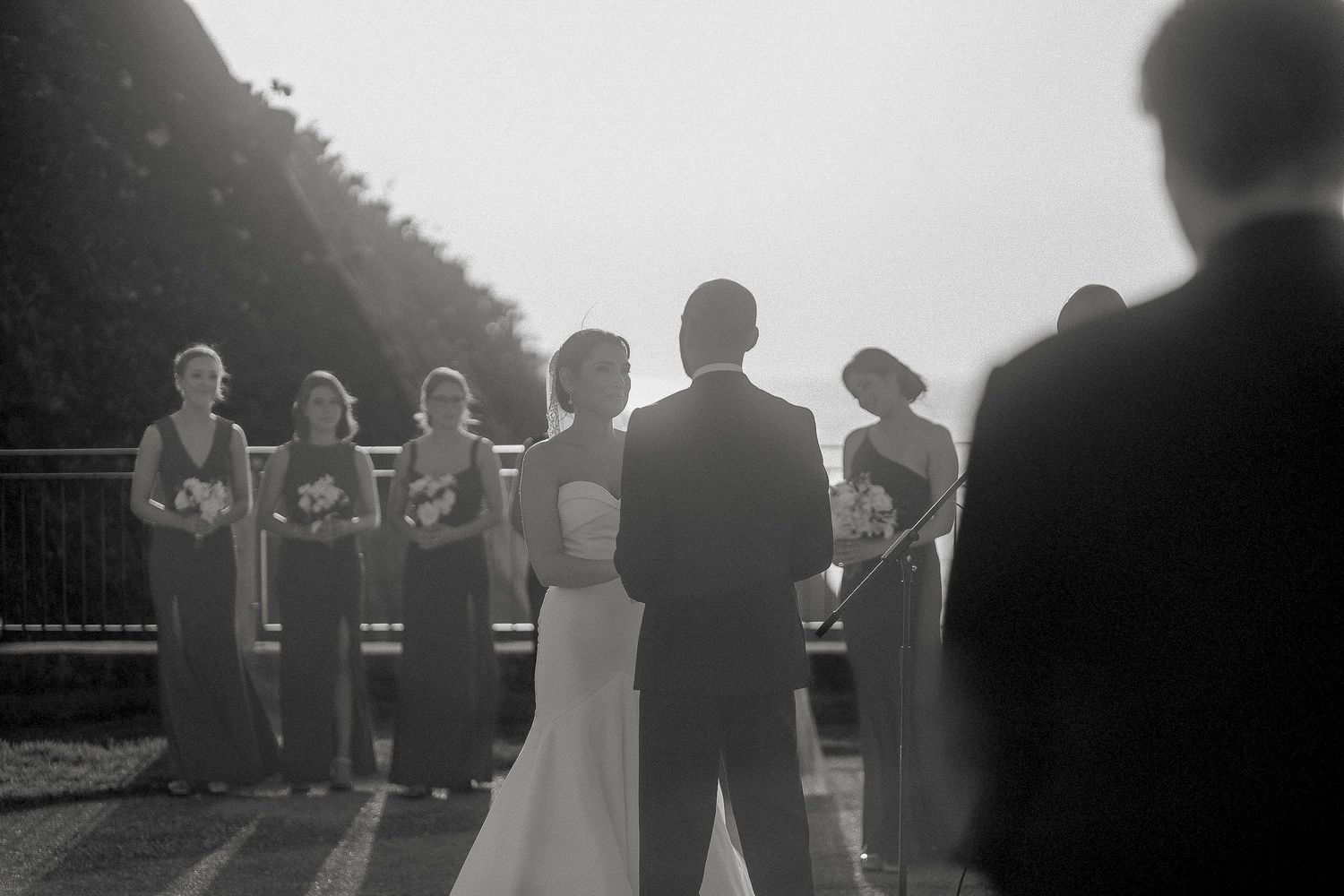 Black and white photo of a bride and groom exchanging vows outdoors with bridesmaids standing nearby.