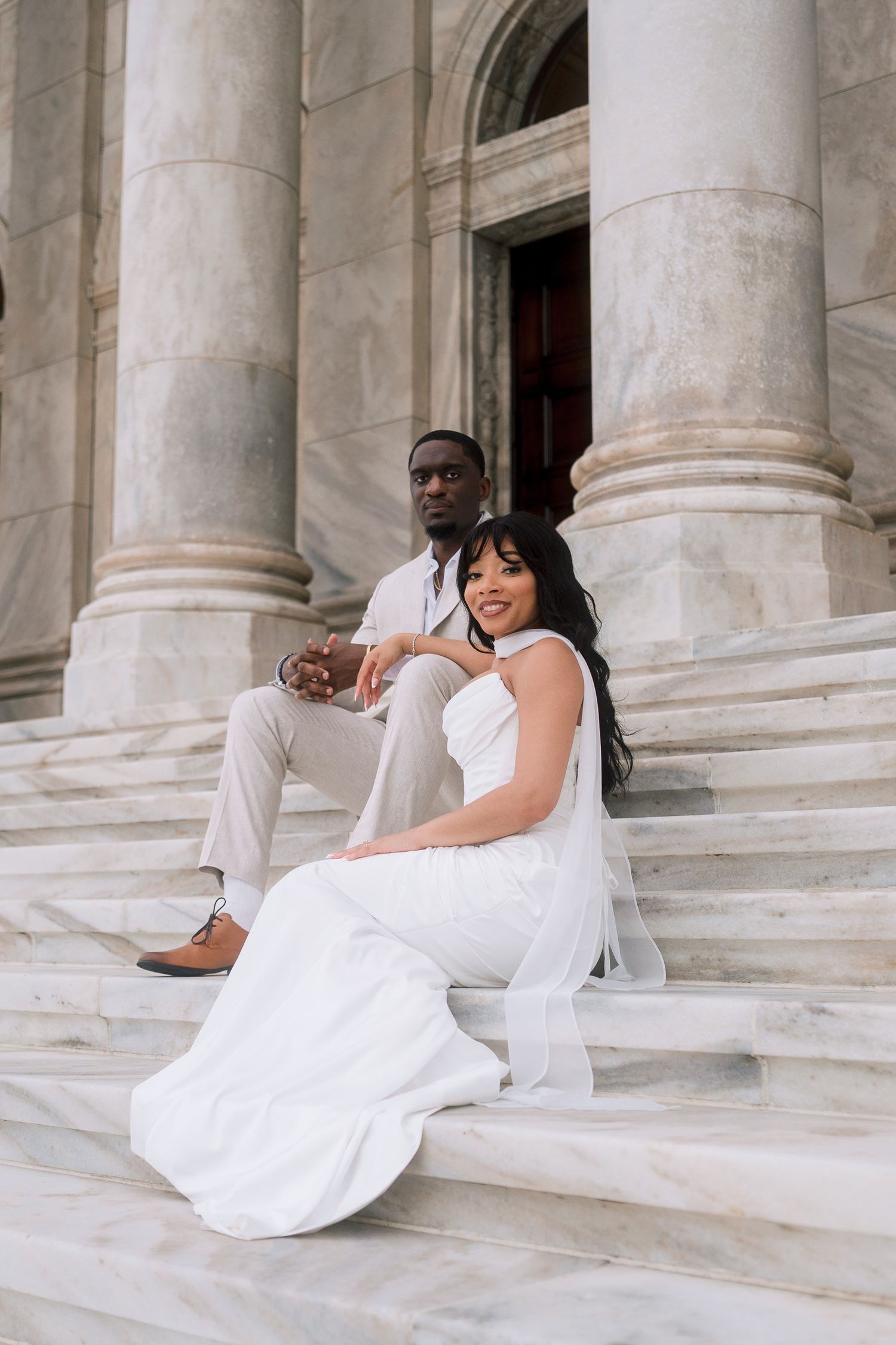 Elegant couple in white attire sitting on marble steps of a grand building for a wedding photo shoot.