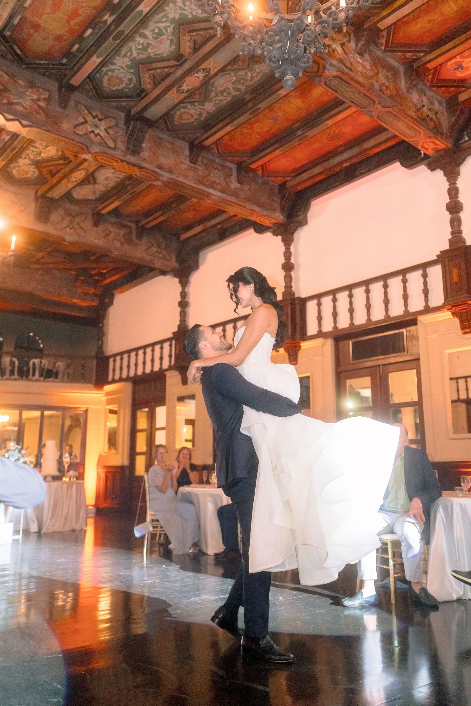 Groom lifting bride in white dress during first dance in elegant ballroom with ornate wooden ceiling.
