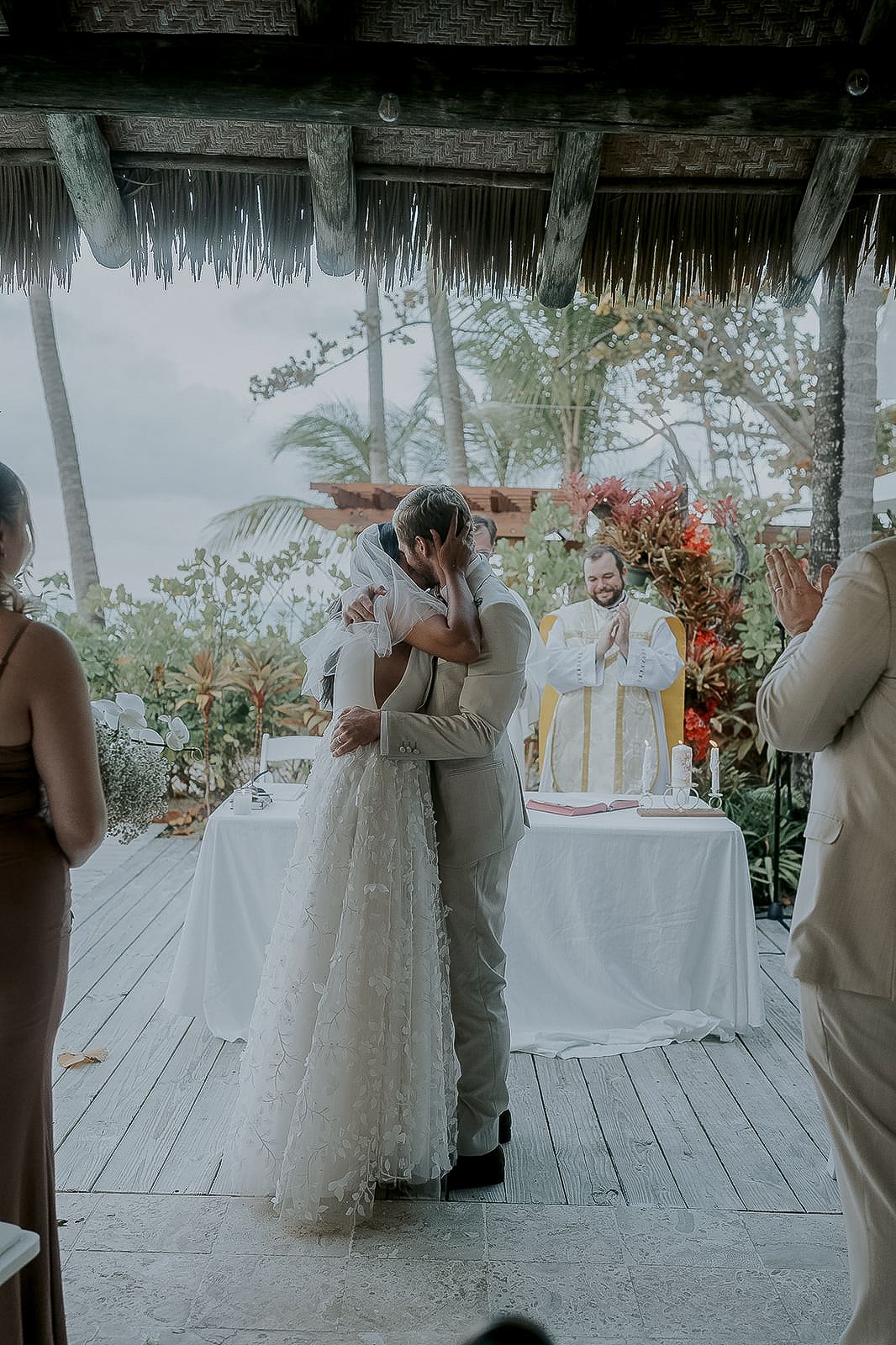 Bride and groom share first kiss at tropical outdoor wedding ceremony with officiant and floral decor.