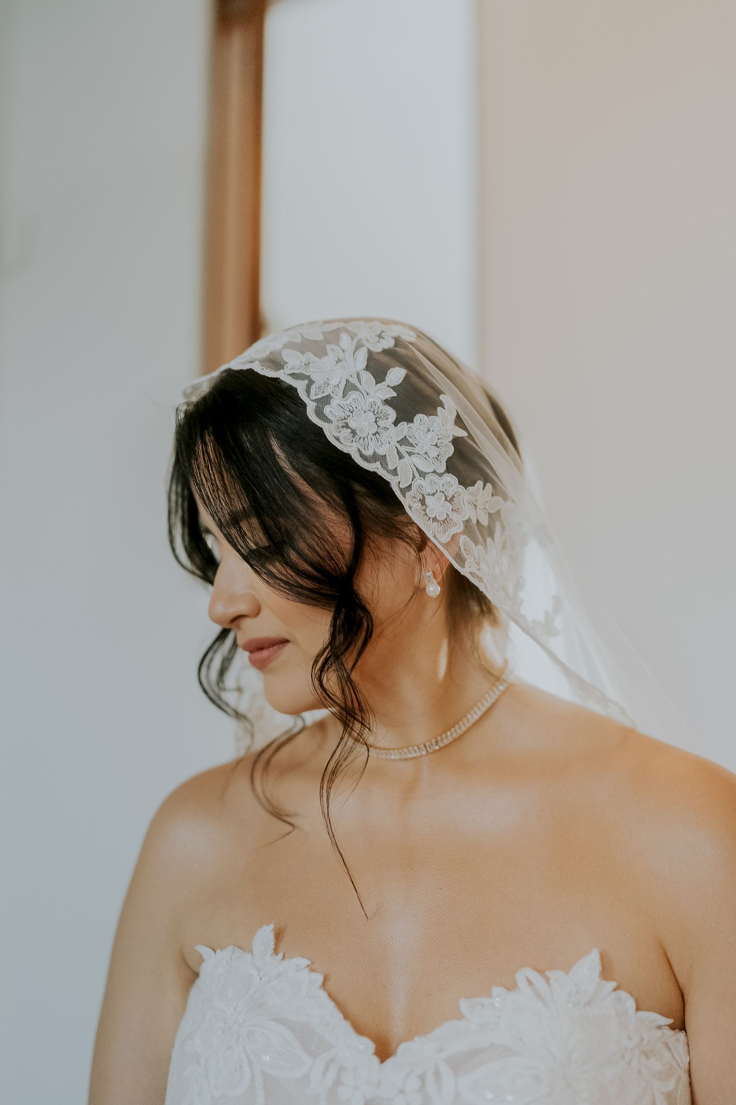 A bride wearing a lace veil and strapless gown with a delicate pearl necklace gazes downward softly.