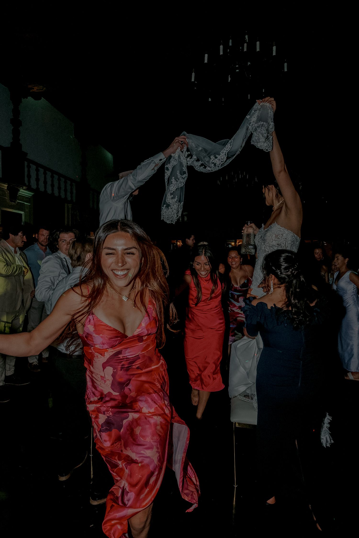 Guest in floral red dress dancing joyfully at lively wedding reception with crowd in background.