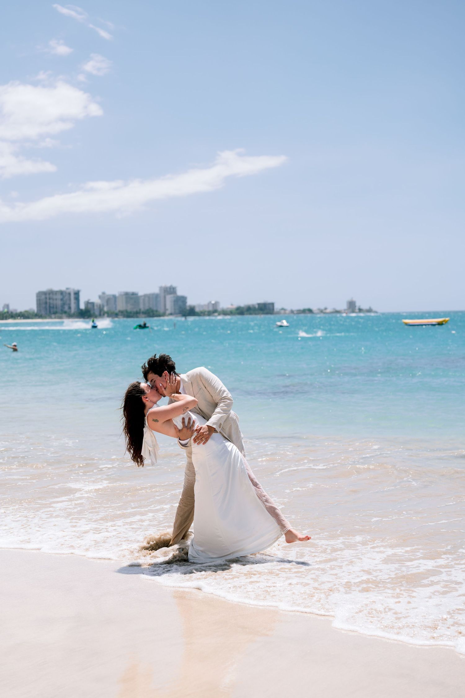 Couple shares romantic kiss on tropical beach shoreline in wedding attire.