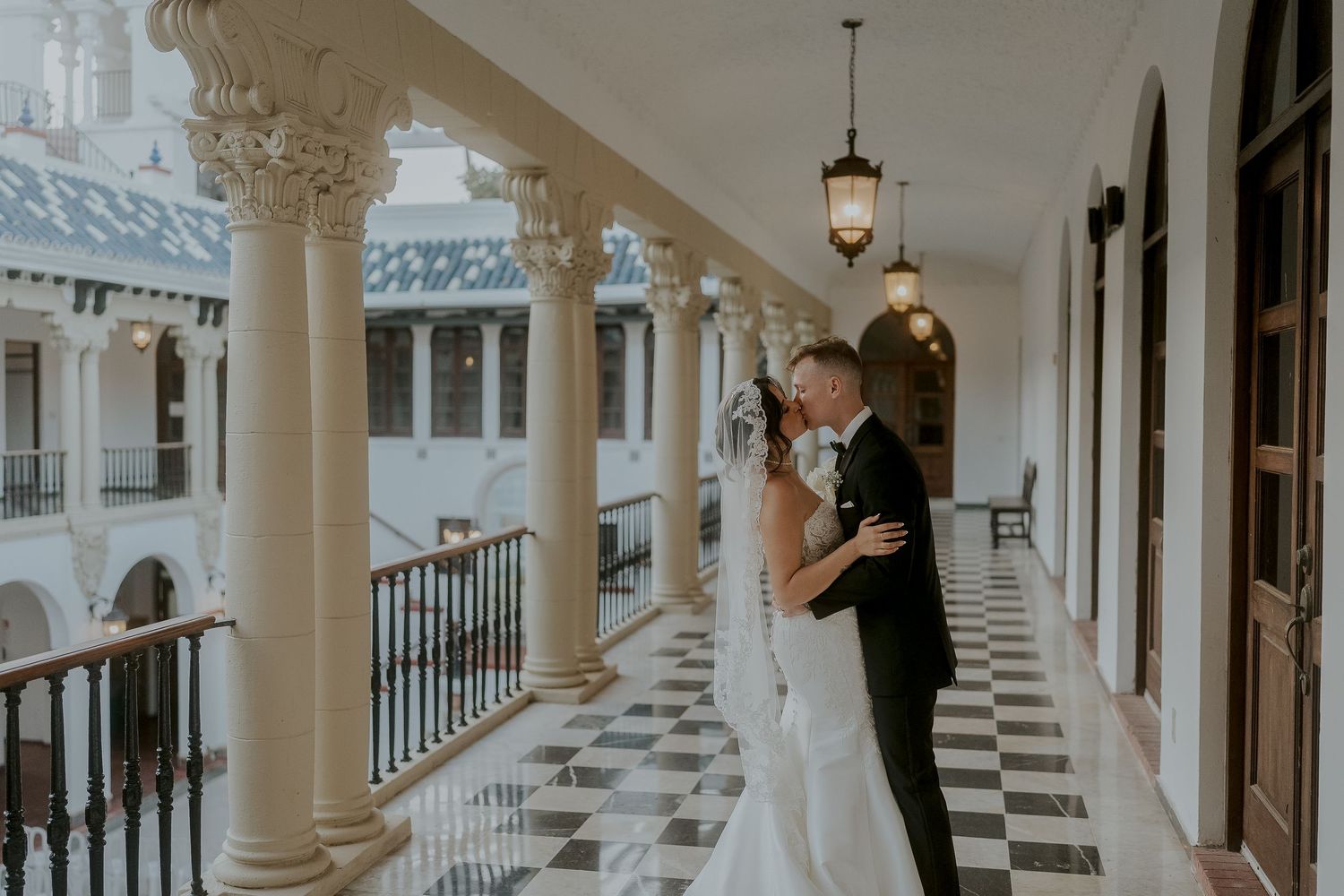 Bride and groom share a romantic embrace on an elegant checkered balcony with white columns and hanging lanterns.