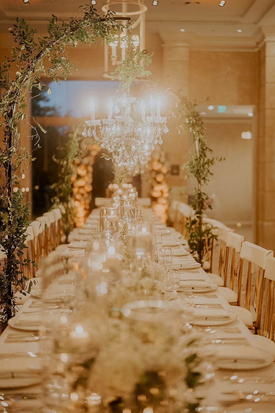 Elegant wedding reception with long dining table, crystal chandelier, greenery decor, candles, and gold chairs in a warmly lit venue.