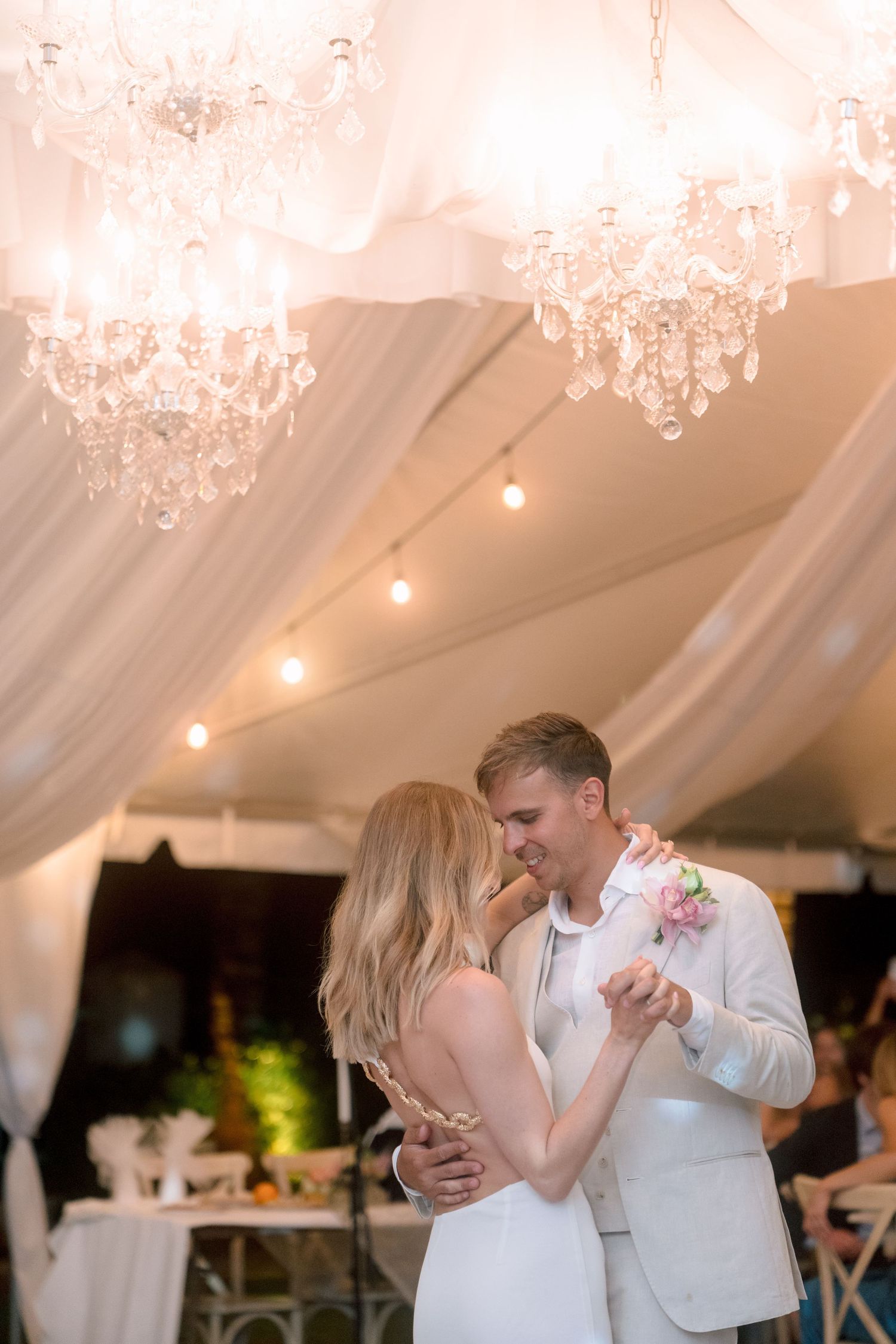 Newlyweds share first dance under elegant crystal chandeliers in a draped white wedding reception tent.