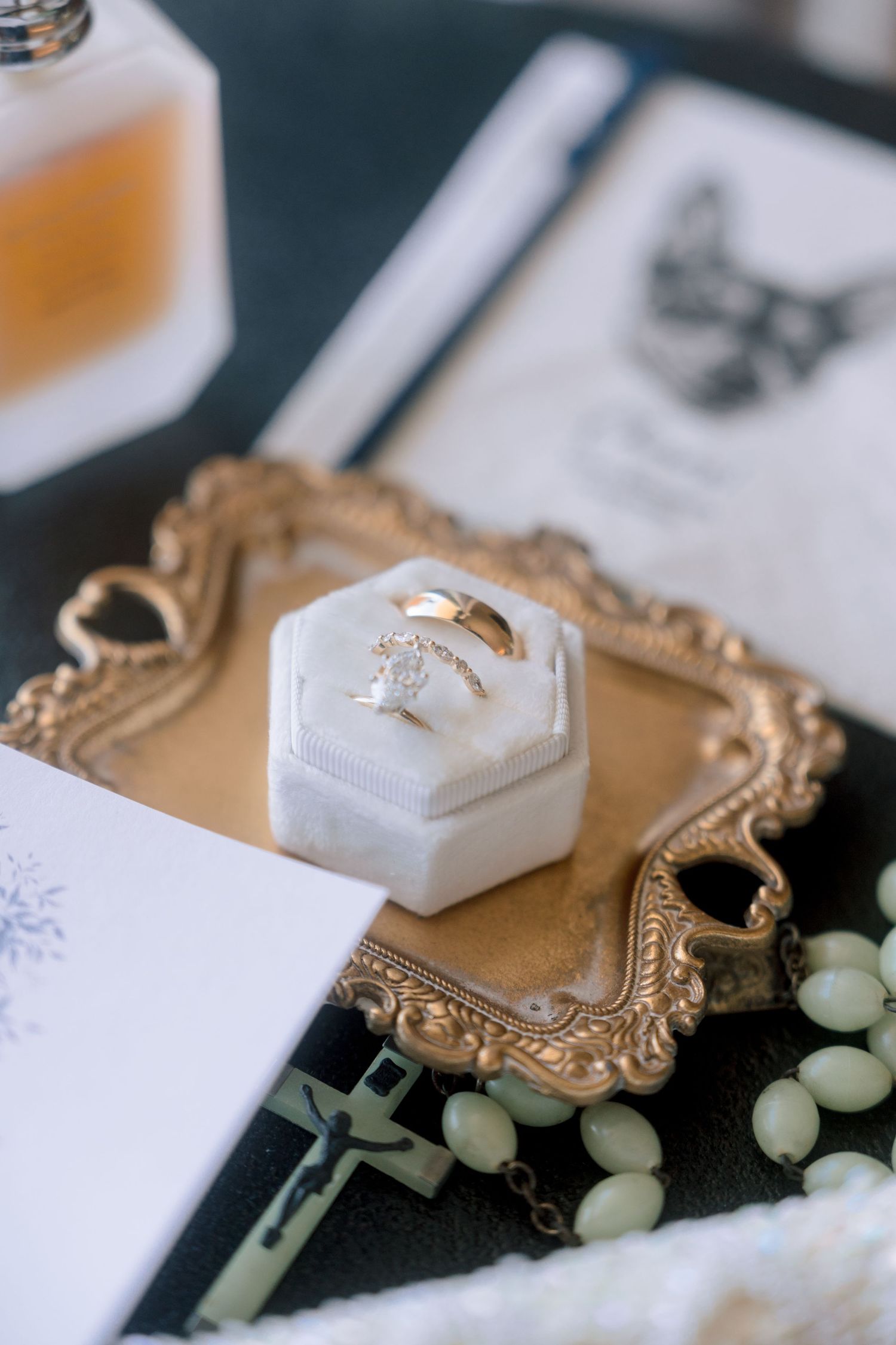 Gold wedding rings displayed in a white velvet hexagon box on an ornate gold tray with rosary beads and perfume.