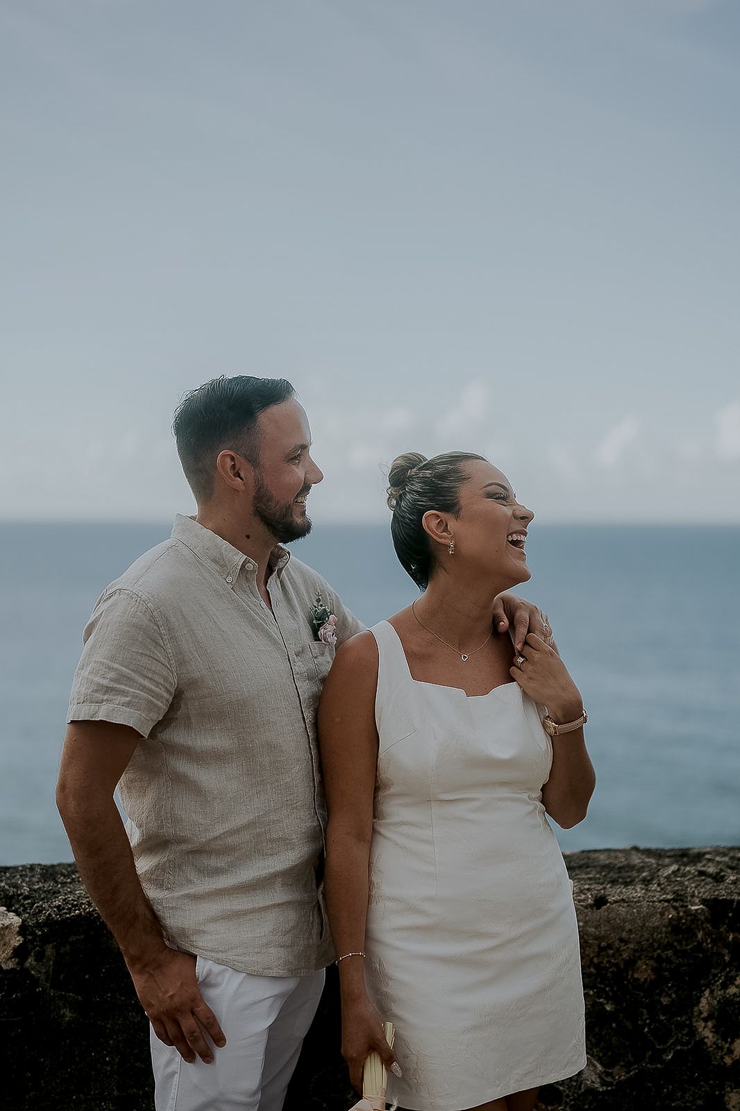 Couple in white attire smiling by ocean wall, beach wedding portrait with blue sea backdrop.
