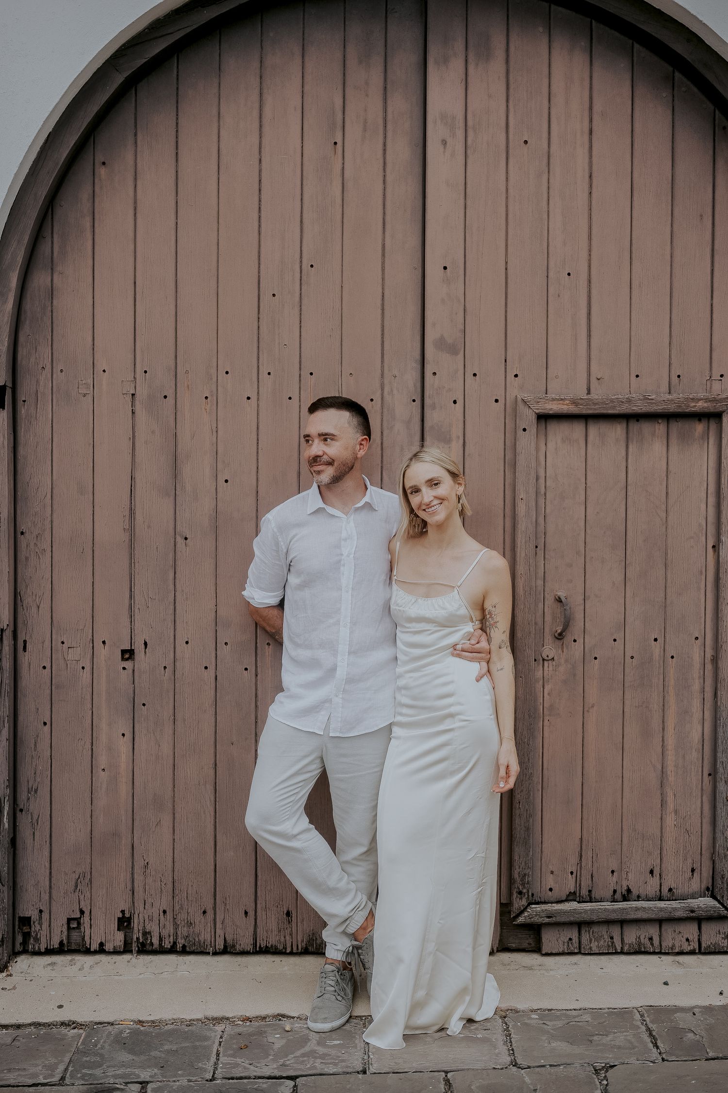 Smiling couple in white outfits posing together against a rustic arched wooden door.
