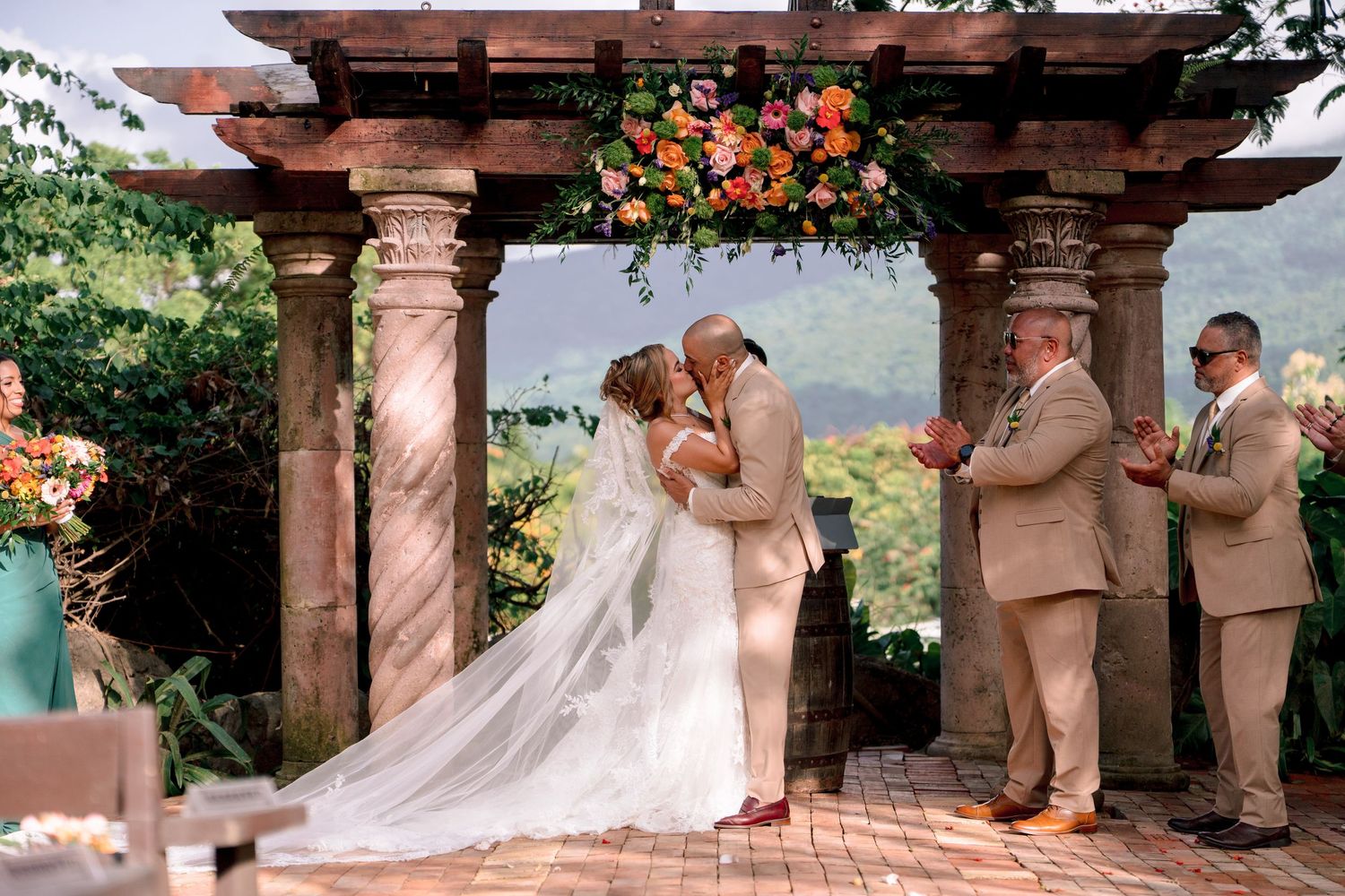 Bride and groom share first kiss under floral pergola at outdoor wedding ceremony with mountain views.