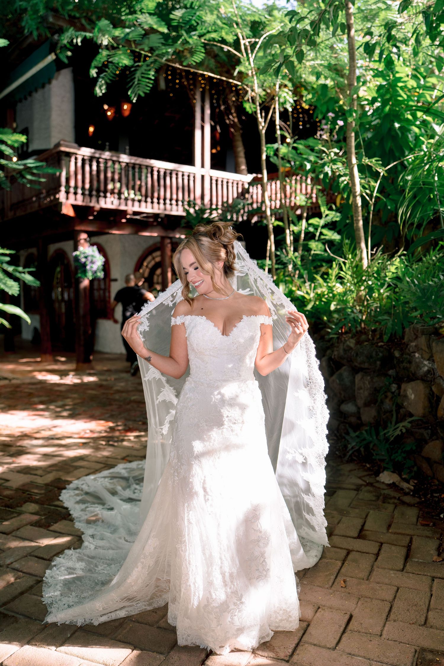 Bride in lace mermaid gown holding flowing veil outdoors near rustic venue surrounded by lush greenery.