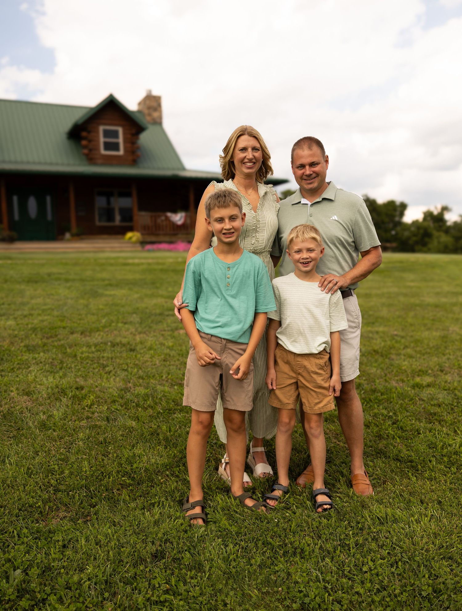 Family of four smiling together on green lawn in front of a rustic log cabin home on a sunny day.