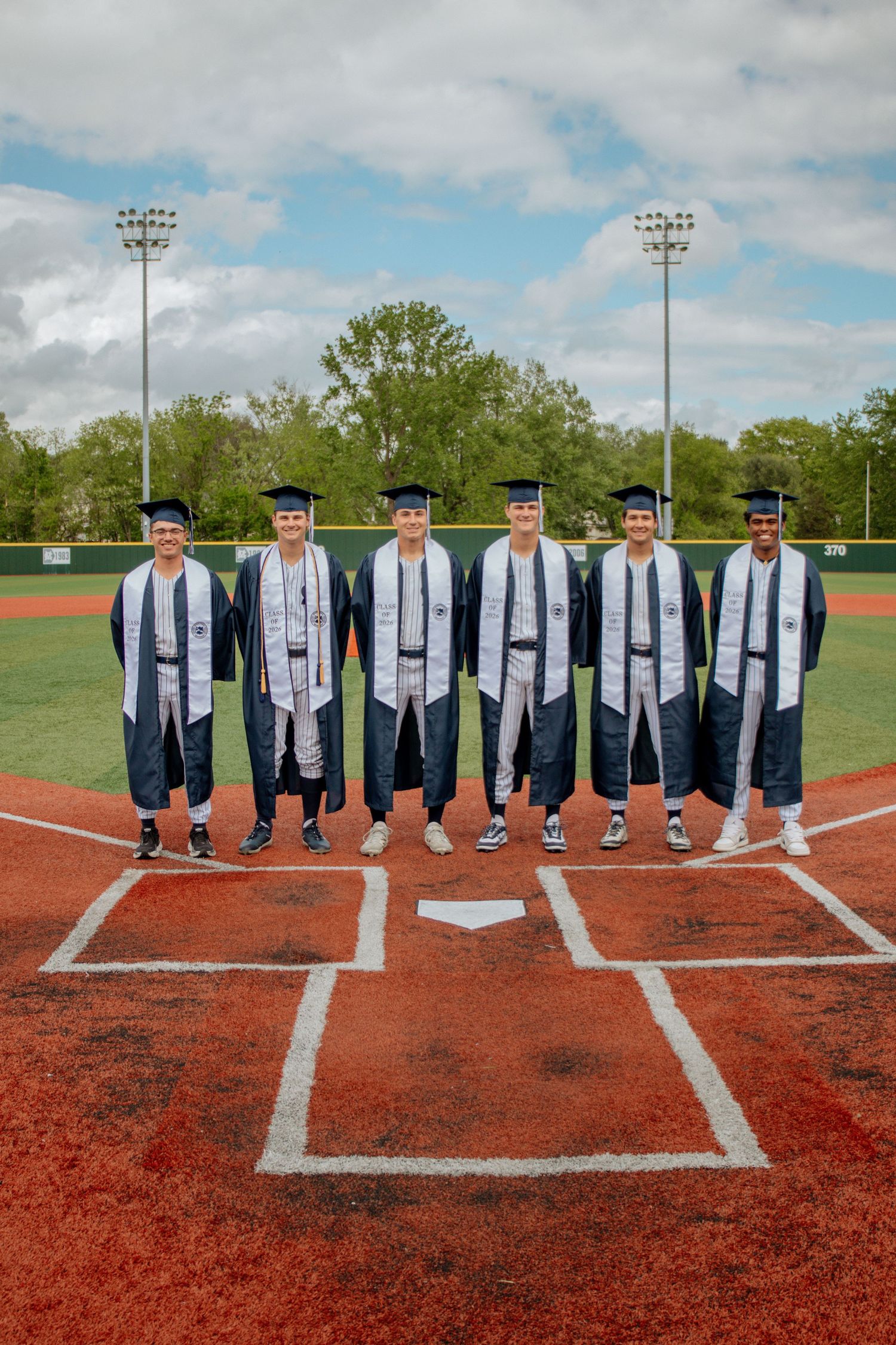 Six graduates in caps and gowns posing at home plate on a baseball field.