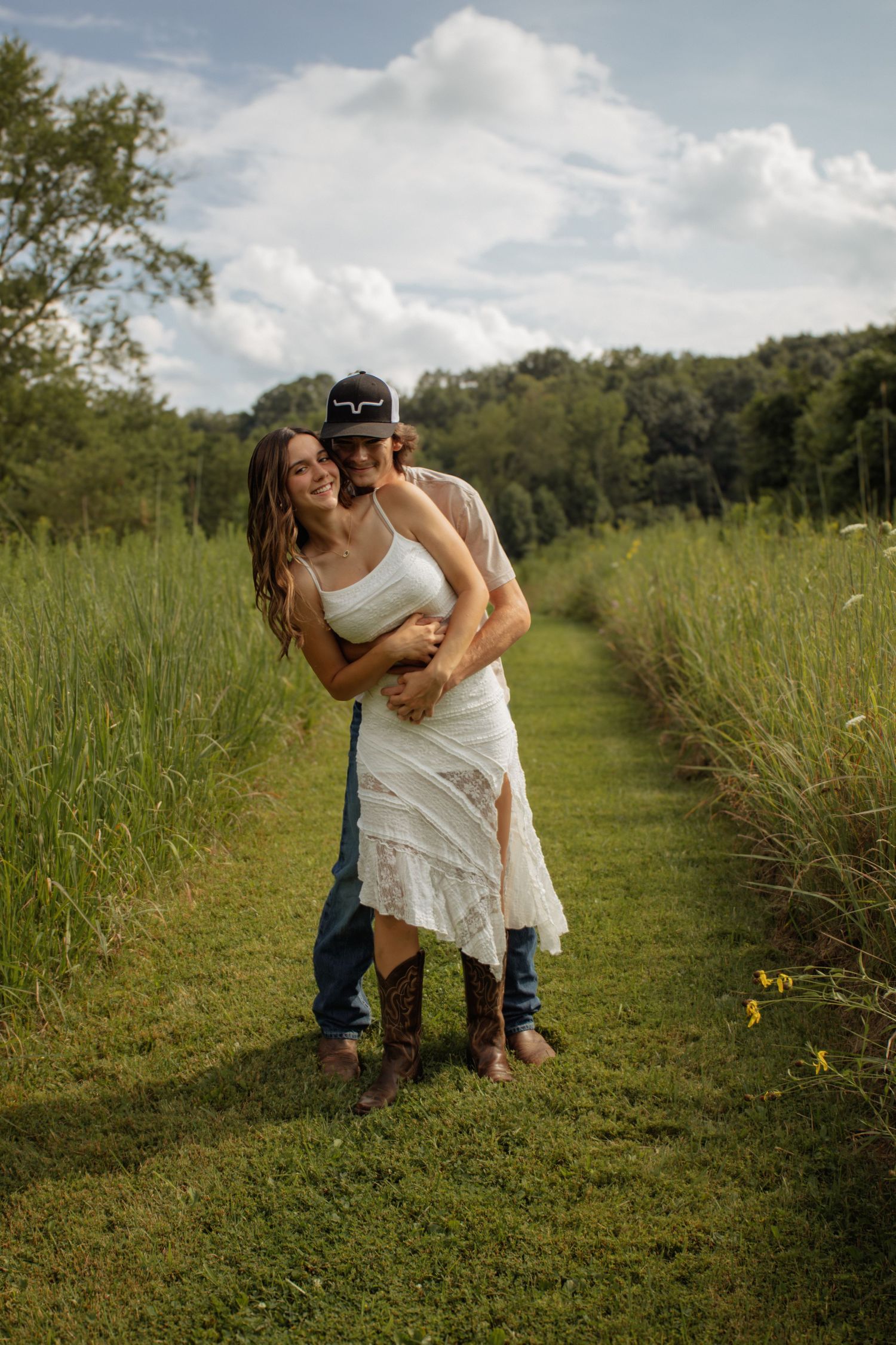 Couple embracing in a green meadow path, woman in white dress and cowboy boots, man in jeans and cap.