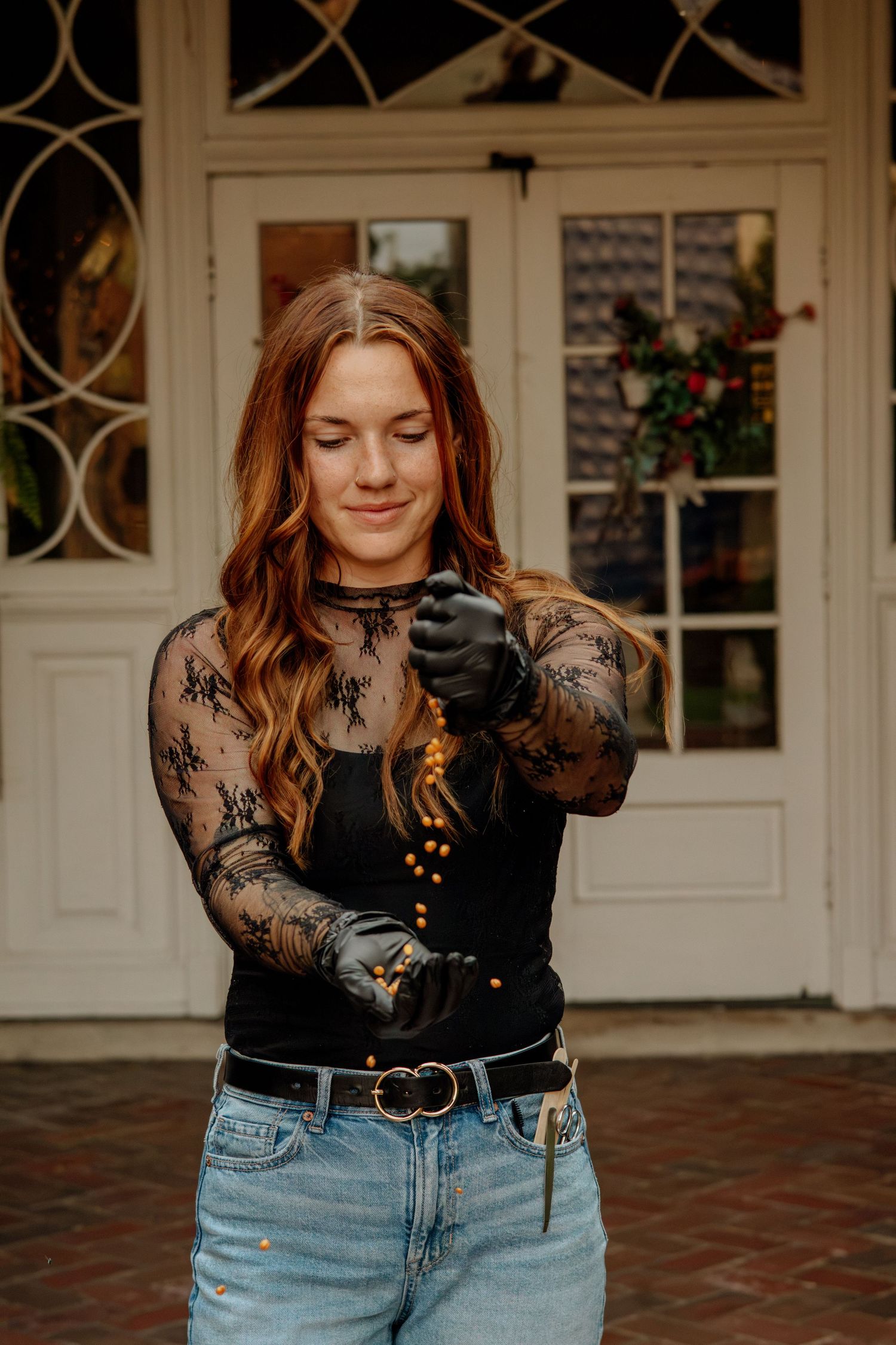 Smiling redhead woman in black lace top and jeans holding gold wax beads outdoors near white door.