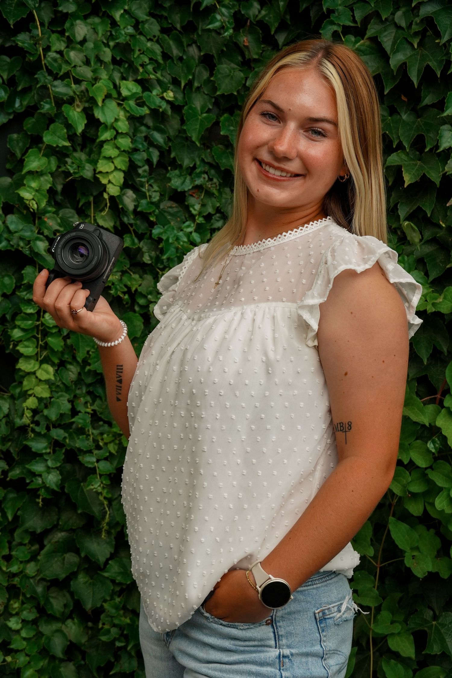 Smiling blonde photographer in white top holds Canon camera against lush green ivy wall.