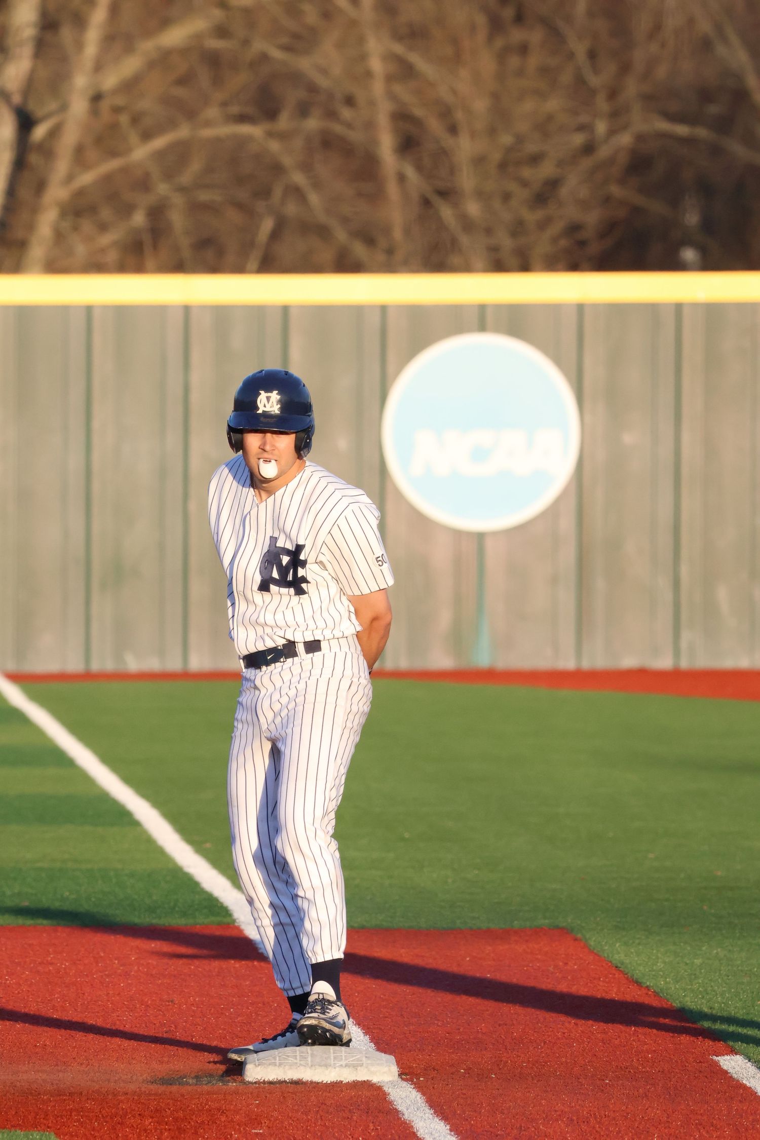 NCAA college baseball player in white pinstripe uniform stands on base during a game at an NCAA-marked field.