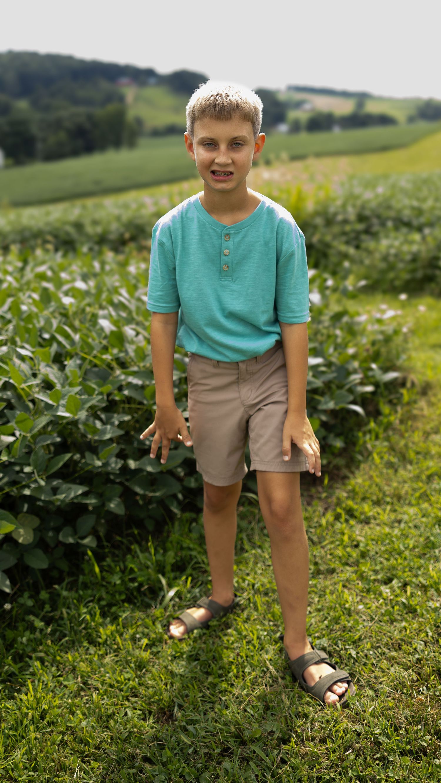 A young blonde boy in a teal shirt and khaki shorts stands smiling in a lush green farm field on a sunny day.