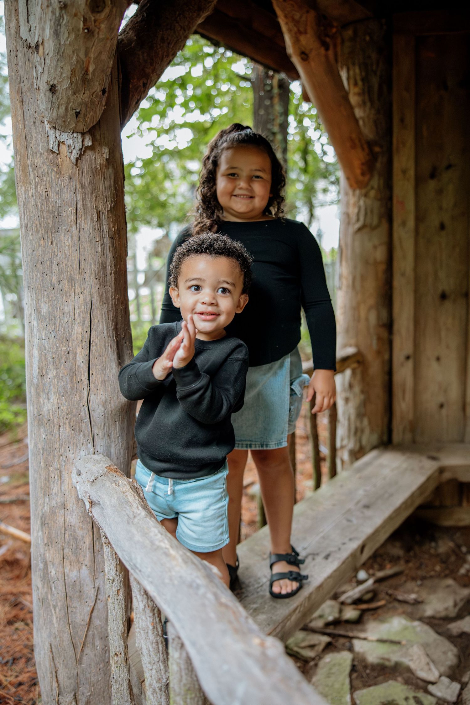 Two young children smiling inside a rustic wooden playhouse structure outdoors.