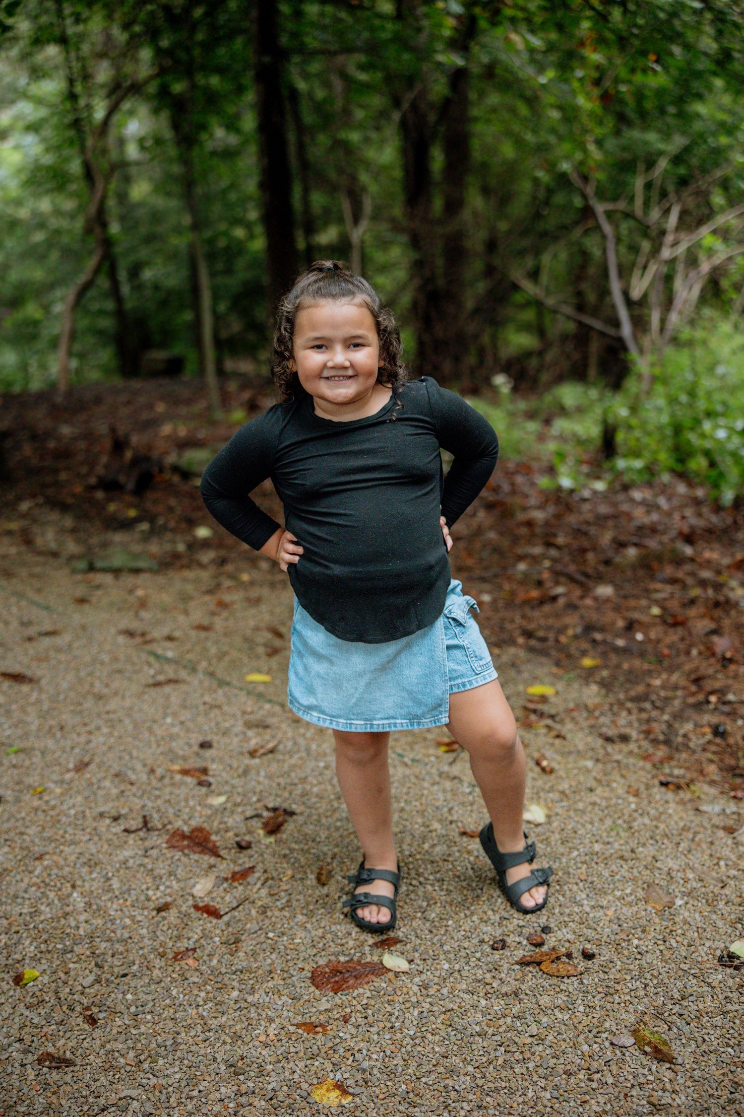 Young girl in black top and denim skirt poses confidently on a forest path, smiling with hands on hips.