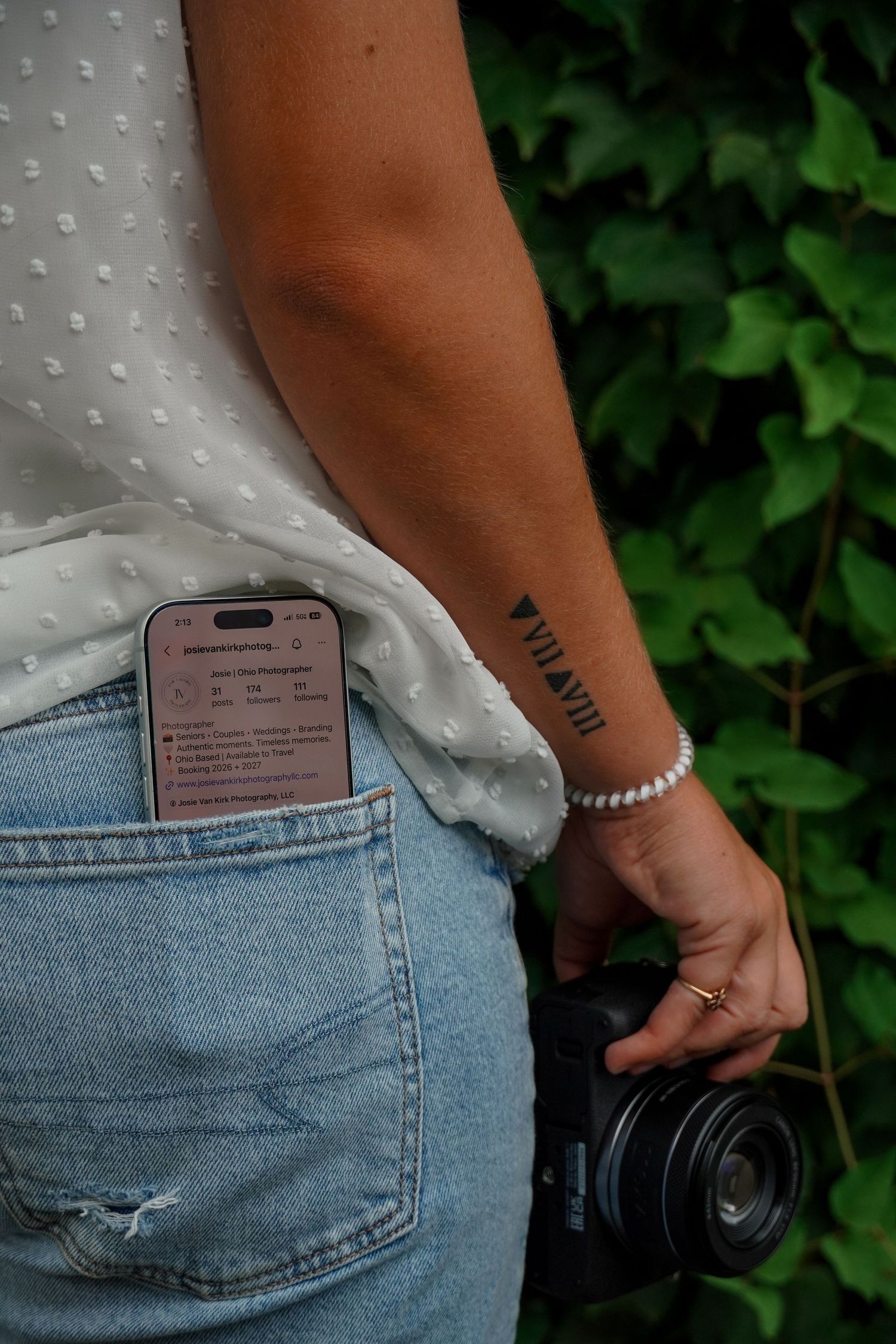 Woman holding camera with wrist tattoo, pearl bracelet, phone in jeans pocket, wearing white blouse outdoors.